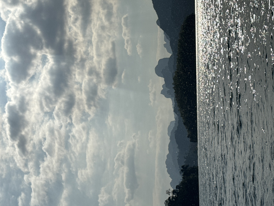 The scene depicts a natural landscape in the city of Unknown, Unknown. The foreground is dominated by a vast body of water, likely a lake or reservoir, exhibiting numerous small ripples and widespread specular reflections from sunlight across its dark, textured surface. In the mid-ground, a long, dark landmass, appearing as a densely forested island or shoreline, stretches horizontally. Behind this, a range of irregular, dark, mountainous formations, consistent with karstic geology, are silhouetted against the brighter sky. The sky, which occupies the upper half of the frame, is heavily covered with a mix of white and light grey cumulus clouds, with visible breaks in the cloud cover in the upper right quadrant from which faint crepuscular rays descend. No human figures, structures, or specific human actions are discernible within the frame.