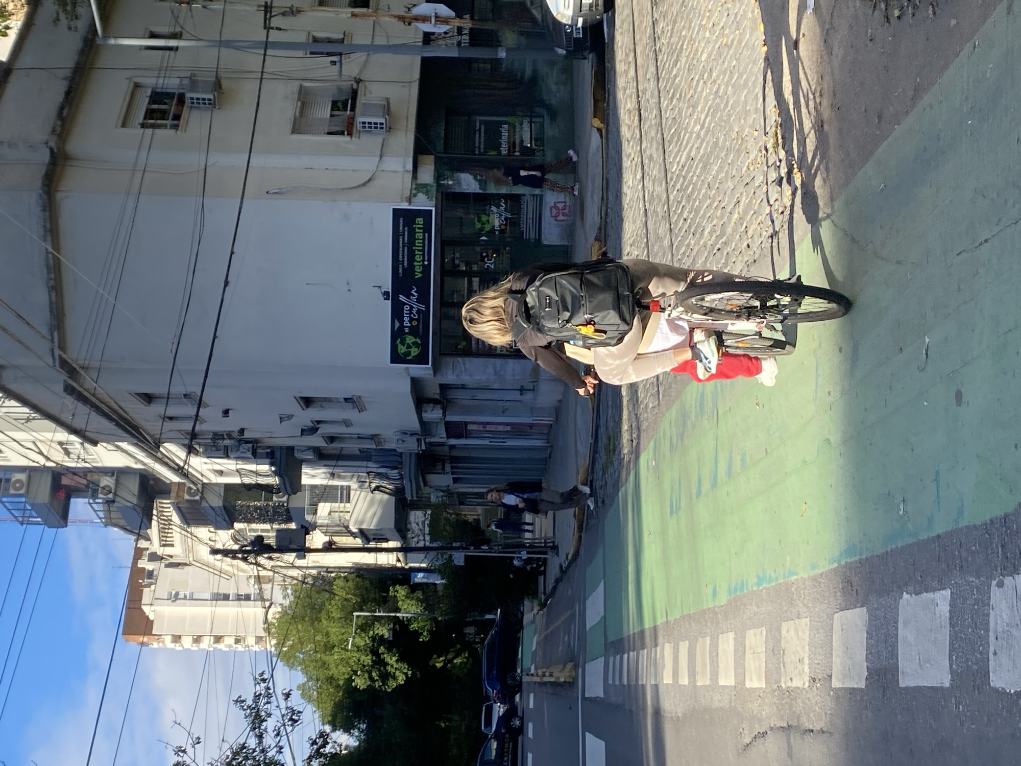A woman rides a bicycle on a green bike lane next to a crosswalk on a city street in Buenos Aires, Argentina. The woman is wearing a red top, beige pants, and a black backpack. A veterinary clinic is visible in the background. Buildings line the street. The sky is partially visible.