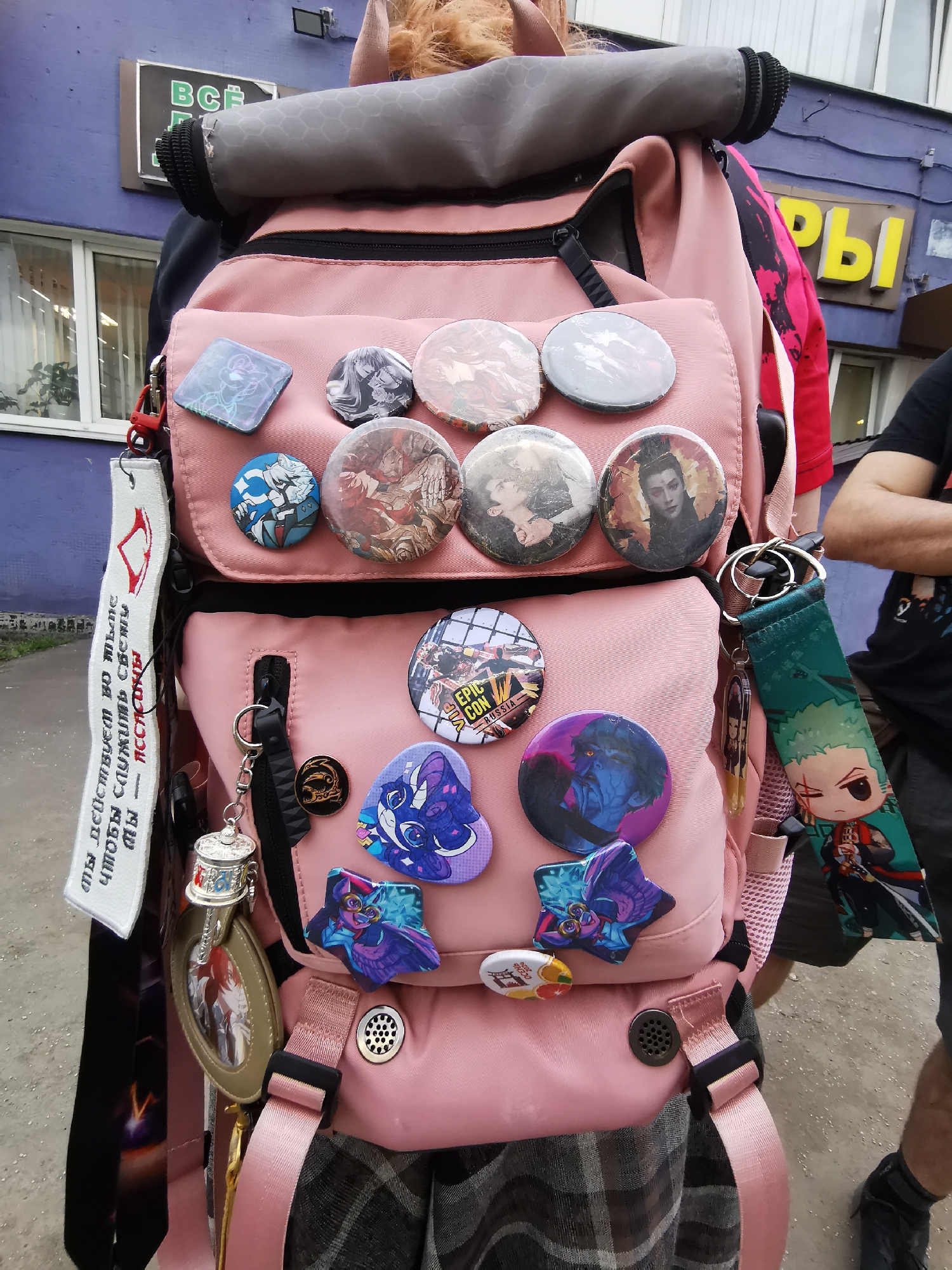 The image shows a person wearing a pink backpack adorned with various pins and keychains. The backpack has two main compartments, each covered with pins. The top compartment features round pins with various character images, while the bottom compartment has pins related to "Epic Con Russia," "My Little Pony," and other character designs. Hanging from the backpack's zippers are keychains with character images and a silver prayer wheel.
A white lanyard with red markings and text written in a Cyrillic script hangs from one side of the backpack. A green lanyard featuring a "One Piece" character is attached to the other side.
The person wearing the backpack is wearing a plaid skirt or pants and a black top. Another person is standing to the right, partially visible with crossed arms. The background shows a blue building with a sign partially obscured, possibly in Russian. The setting appears to be outdoors in a city, with a sidewalk visible.