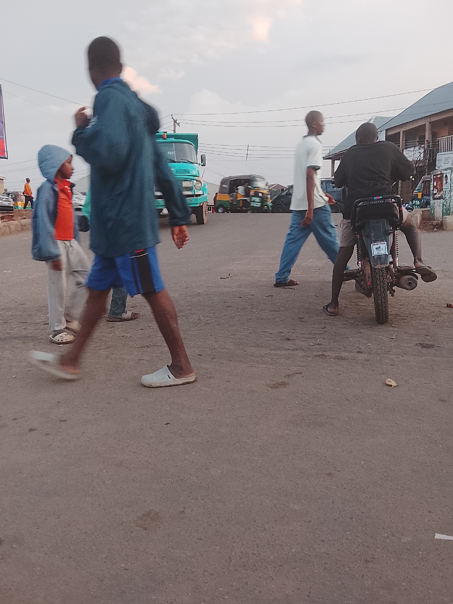 In Gwong, Nigeria, the scene captures a street intersection. Four individuals are present: two are pedestrians – one wearing a blue hooded jacket over an orange shirt with grey pants and the other in a blue jacket and shorts – one is walking away in a white shirt and jeans, and one sits atop a parked motorcycle in a black shirt. In the background, a green truck is parked on the side of the road, while auto rickshaws are visible in the mid-ground. A two-story building with a dark roof is on the right.