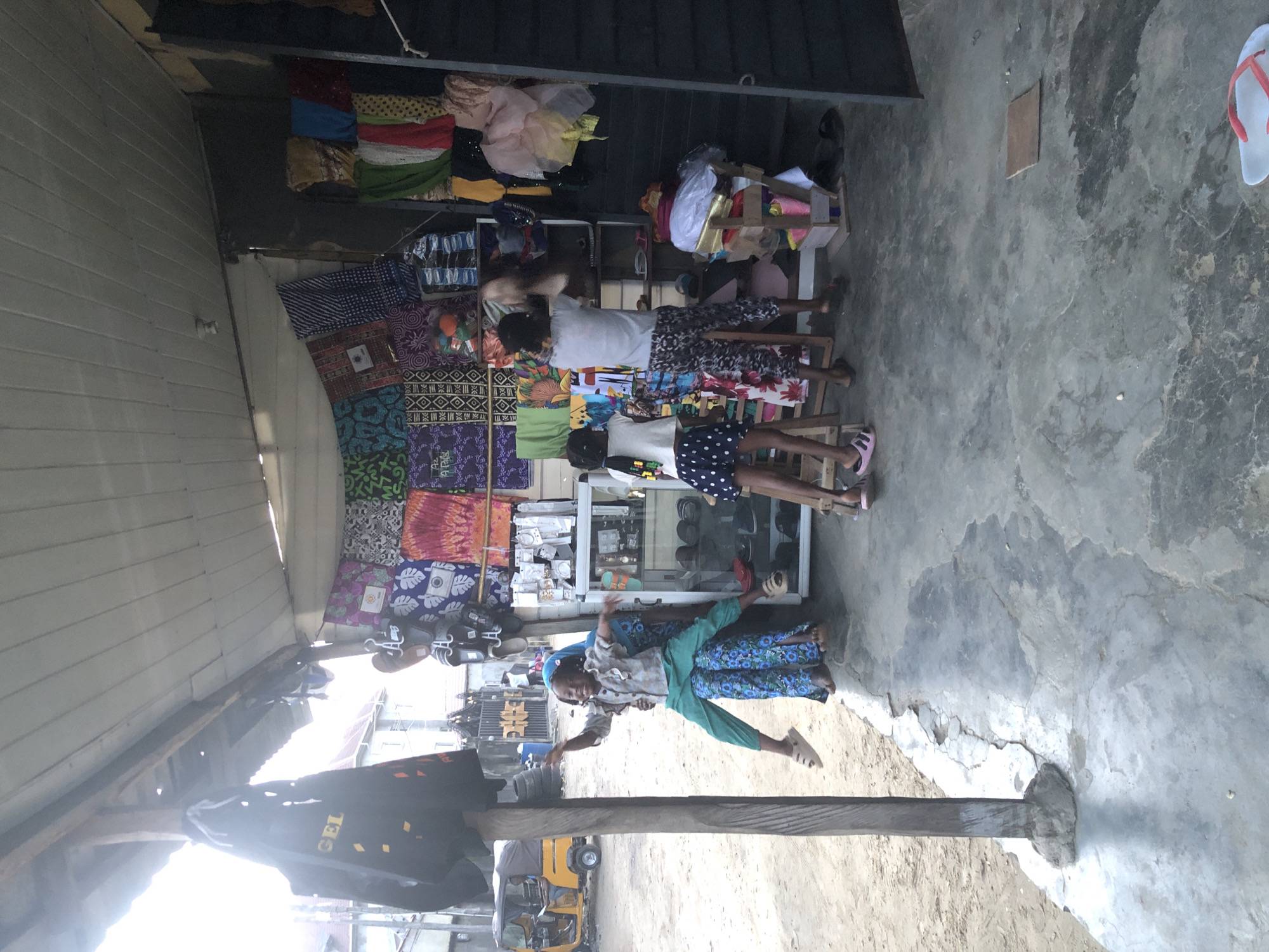 In a marketplace stall in Unknown, Unknown, four children are engaged in playful activity. One child, on the left, is being held by another, both appearing to be enjoying themselves. To the right, two other children are interacting within the stall. The stall is filled with colorful textiles, footwear, and various merchandise displayed on shelves and hanging racks. The ground is a concrete surface with some wear and tear, and the stall is covered by a corrugated metal roof. In the background, a yellow auto-rickshaw is visible on a dirt road, suggesting an outdoor setting.