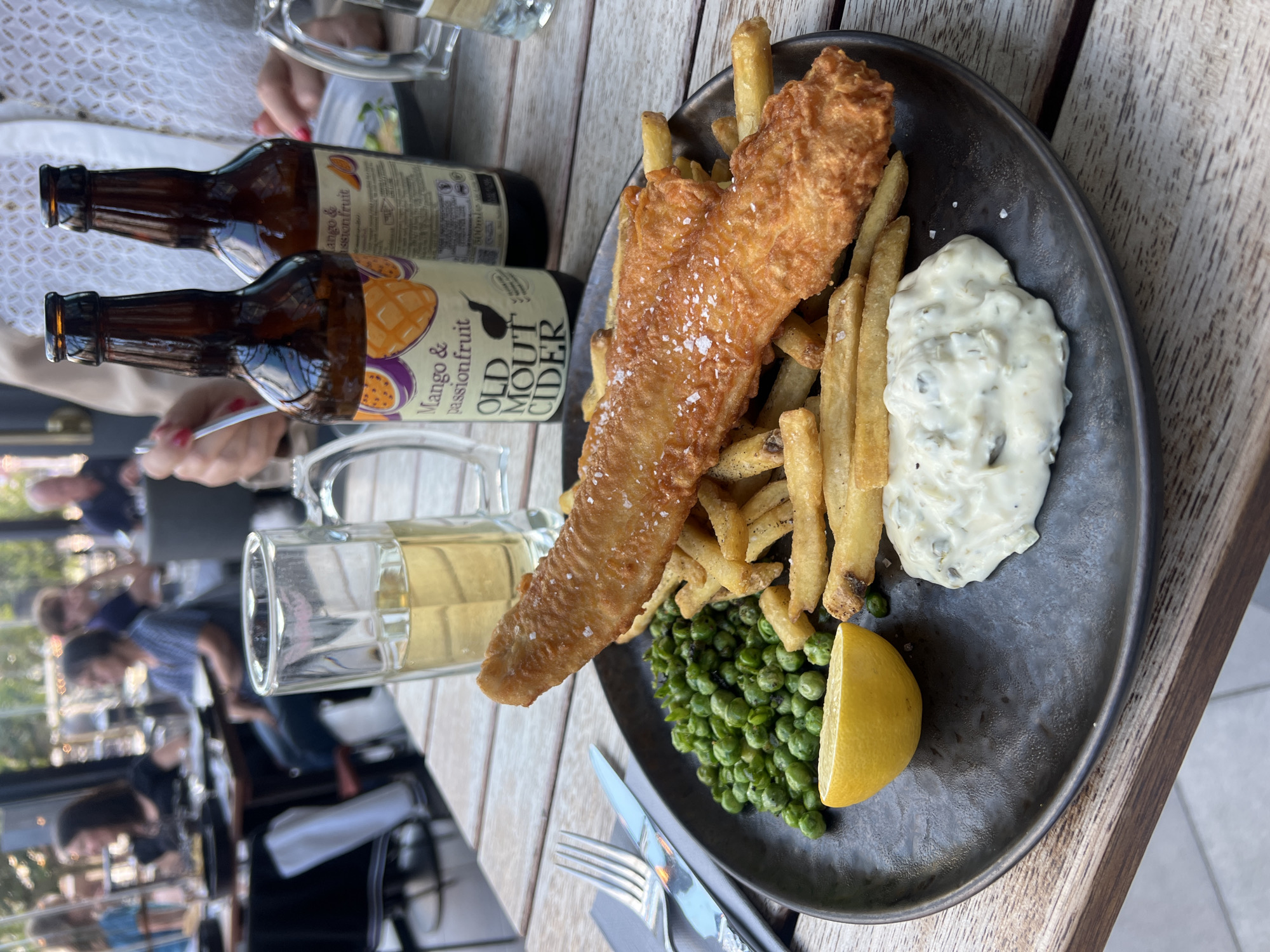 This is a close-up, eye-level shot of a plate of fish and chips served at a restaurant, likely located in Portsmouth, United Kingdom, given the presence of "Old Mout Cider" which is popular in the UK. The food is presented on a dark, textured plate. The main subject is a large piece of golden-brown, battered fish resting on a bed of thick-cut french fries. Scattered peas and a lemon wedge are visible to the left of the fish and chips, and a dollop of tartar sauce is to the right. The meal is accompanied by a tall glass of pale ale or cider and two bottles of "Old Mout Cider" labeled "Mango & Passionfruit." The wooden table surface is visible in the foreground and background, suggesting an outdoor or semi-outdoor dining area with people seated at other tables in the blurred background.