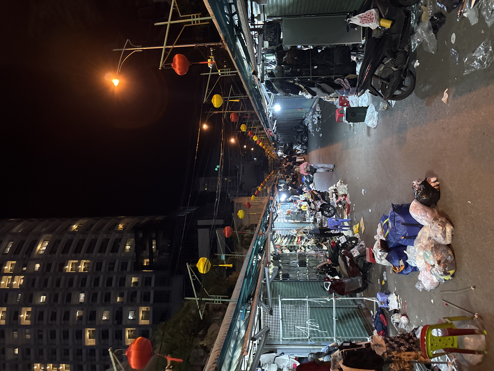 The image depicts a street market at night in Nha Trang Ward, Vietnam, with a large, modern apartment building looming in the background. The market stalls are lined up along a paved pathway, and the scene is illuminated by streetlights and red and yellow lanterns hanging overhead.
In the foreground, the pathway is littered with trash, including plastic bags, debris, and discarded items. Several motorcycles are parked along the sides of the market. Various goods are displayed and piled in front of the stalls, with some items hanging on racks and others in large bags.
A few people are visible in the market. In the mid-ground, a woman in a pink shirt and light-colored pants is walking away from the camera. Further back, several other individuals can be seen browsing or working at the stalls.
The apartment building in the background is illuminated with many lights, suggesting it is inhabited. The overall atmosphere is that of a busy, albeit somewhat messy, nighttime market.