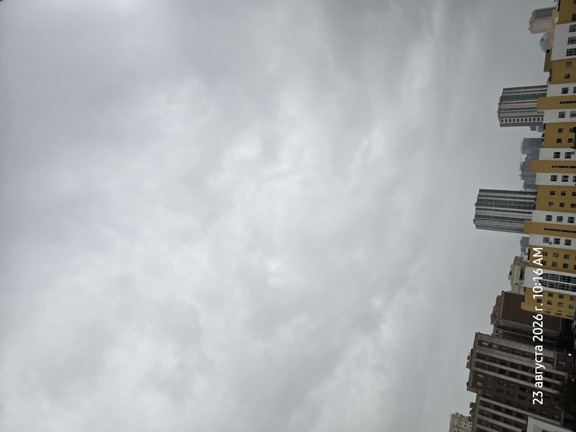 The image shows a cityscape under a cloudy sky. In the foreground, the text "23 августа 2026 г. 10:16 AM" is superimposed, indicating the date and time. Several multi-story buildings are visible, varying in architectural style and color. Some buildings are predominantly brown and beige, featuring numerous windows. Other buildings are brightly colored yellow and white, also with many windows. Taller, more modern skyscrapers with green and white striped facades and a building with a distinctive dome are also present. The sky is overcast with dense gray clouds, suggesting a potentially overcast or rainy day. The scene is set in the city of Astana, Kazakhstan. There are no people visible in the image.