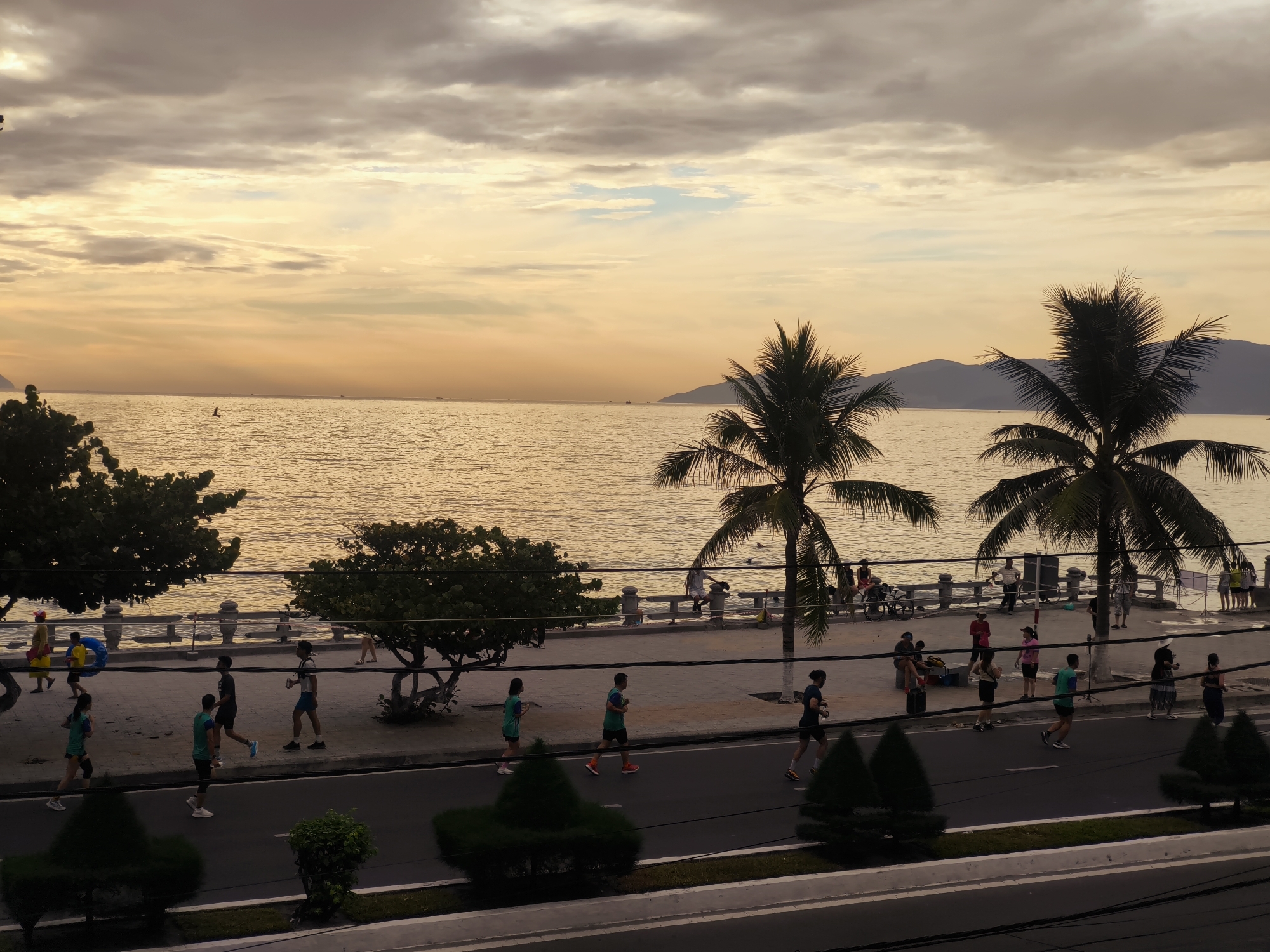 A group of people are jogging on a paved road beside a walkway along the coast of Nha Trang, Vietnam. The sun is low in the sky, casting a warm glow over the water and clouds. Palm trees line the walkway, and in the distance, mountains can be seen. The scene appears to be a popular spot for morning or evening exercise.