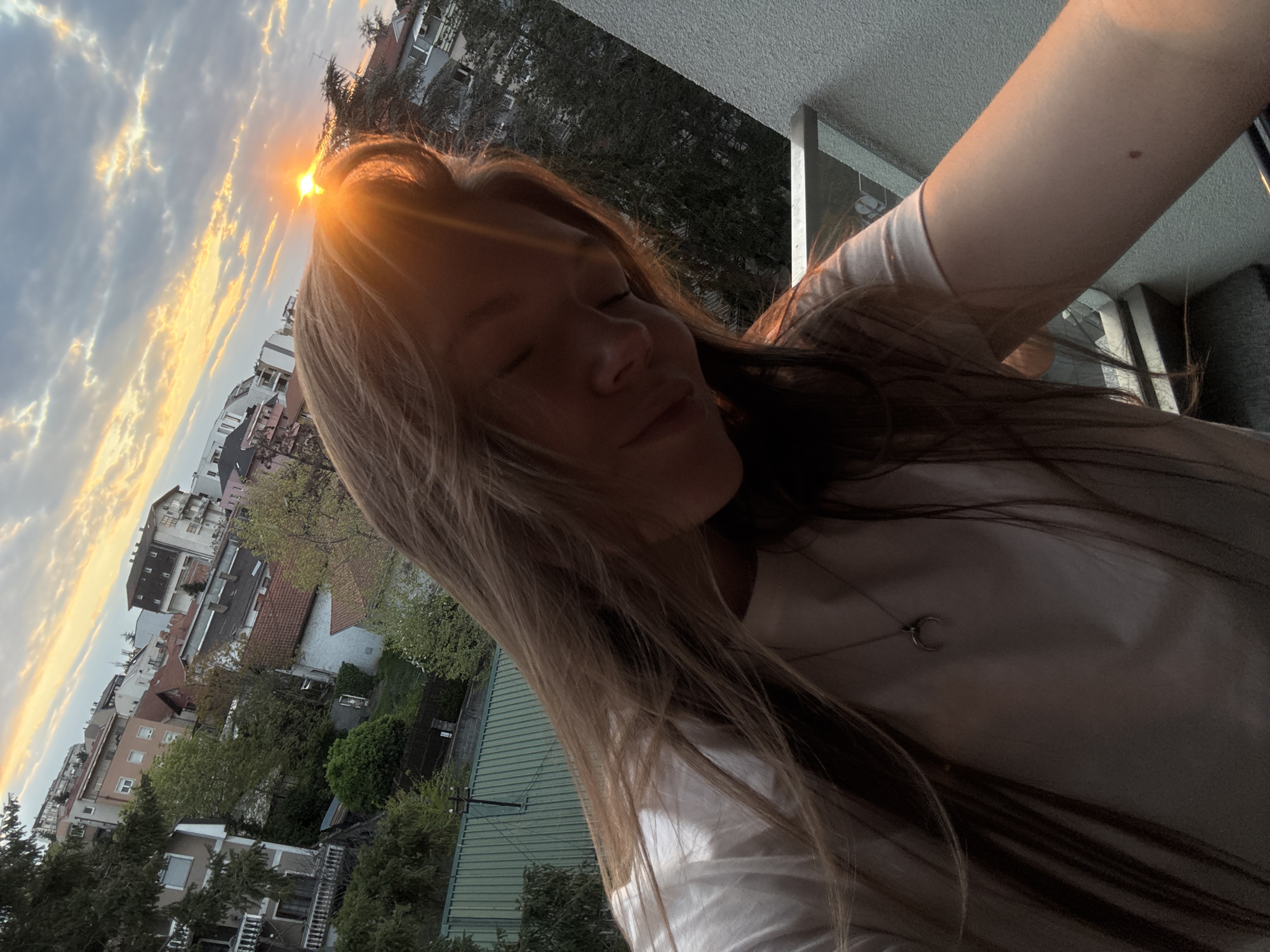 The image captures a dynamic outdoor selfie, taken at an oblique angle, featuring a young person, likely female, with long, light brown hair. The subject appears to be standing on a balcony or elevated outdoor space during either sunrise or sunset, with the sun prominently positioned behind her head. The low sun creates a strong lens flare and bathes her hair in a golden glow, while her eyes are closed or squinting, conveying a sense of peace and enjoyment of the warm light. She is wearing a light-colored t-shirt, and a delicate silver necklace with a crescent moon pendant hangs on her chest. Her arm, extended to take the photo, is partially visible in the foreground.
The background showcases a dense urban or suburban residential area, characteristic of Vozdovac Urban Municipality, Serbia. Multiple multi-story buildings with various architectural designs and tiled roofs are visible, interspersed with abundant green trees and foliage. The sky is partly cloudy, with the clouds illuminated by the warm, golden and orange hues of the rising or setting sun. No visible text is present in the image.