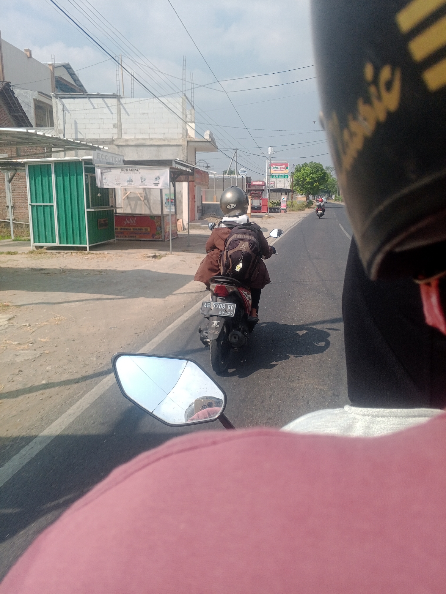 The image depicts a street scene in Pintu, Indonesia, on a sunny day. The perspective is from the back of a motorcycle, looking forward. In the foreground, the back of a rider's head and shoulders, wearing a helmet and a pink shirt, are visible. A side mirror of the motorcycle shows a reflection of another person holding a phone.

Further ahead on the road, another motorcycle is seen from behind, with a rider wearing a brown jacket and a helmet. The motorcycle has a license plate that reads "AE 706 GF".

On the left side of the road, there are several buildings and structures. One building appears to be under construction with exposed brickwork and metal beams. Next to it, there are two green stall-like structures. Further down the road, a sign with the text "RIZKY MART" is visible, along with other commercial establishments. Trees are scattered along the roadside. The sky is clear and blue with some scattered clouds. The road surface is asphalt and appears to be in good condition.