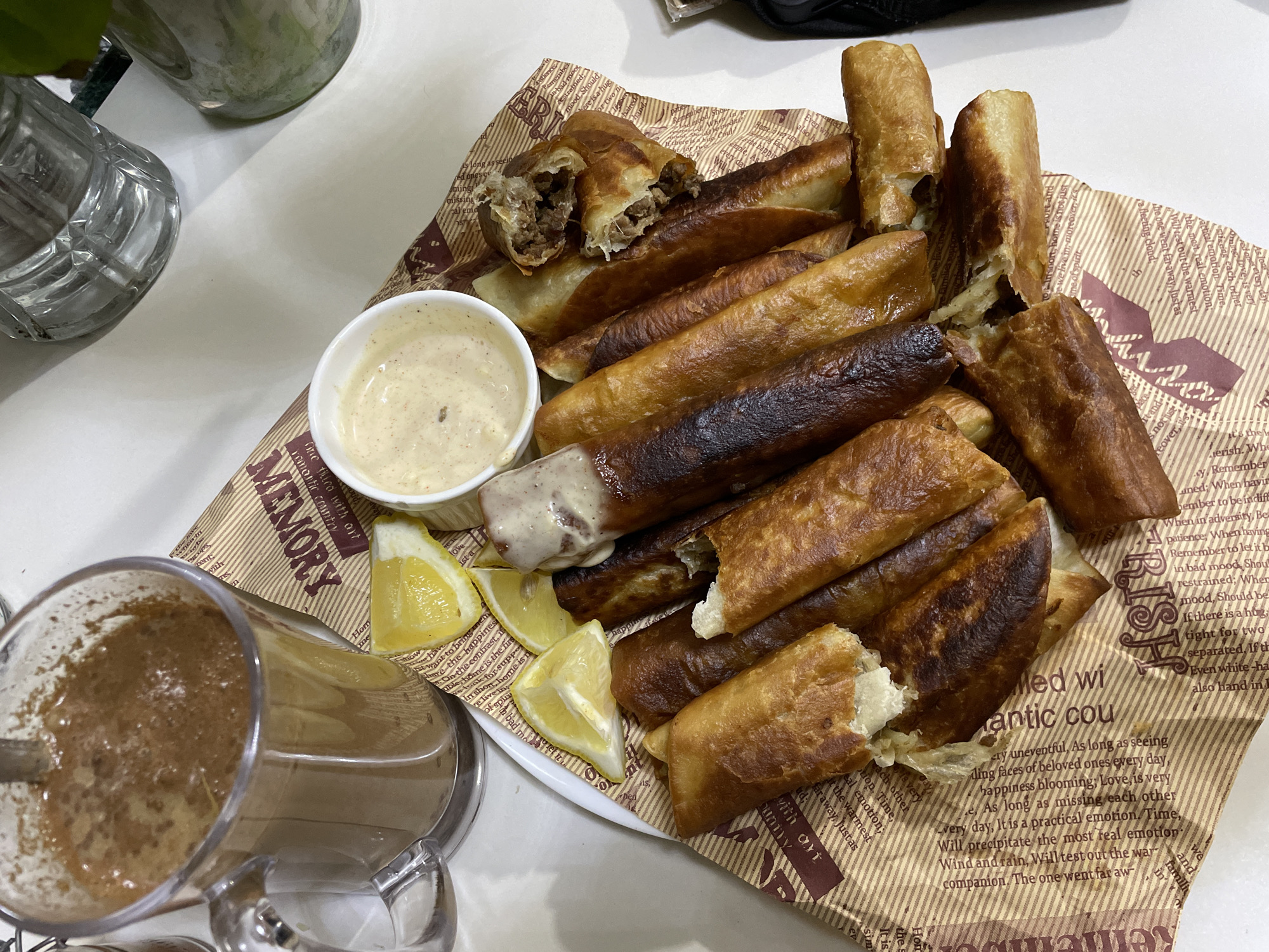 The image shows a plate of fried lumpia rolls arranged on a piece of printed paper with "MEMORY" and other words printed on it. A white bowl with a light-colored sauce and a few lemon wedges are also on the plate. A glass pitcher containing a brown beverage is visible on the side. The setting appears to be a table in a dining establishment. While it is impossible to confirm the location in Davao City, Philippines, based solely on the image, the food presentation is consistent with local cuisine. The lumpia rolls appear to be stuffed with either meat or vegetables. Some rolls are partially broken open.