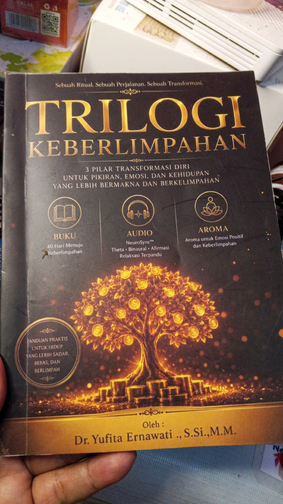 The image is a close-up, overhead shot of a book titled "TRILOGI KEBERLIMPAHAN" which translates to "Trilogy of Abundance." The book cover features a prominent illustration of a tree laden with gold coins, with stacks of coins at its base, symbolizing wealth and prosperity. Above the title, smaller text reads "Sebuah Ritual. Sebuah Perjalanan. Sebuah Transformasi." (A Ritual. A Journey. A Transformation.). Below the main title, there are three pillars of self-transformation: "BUKU" (Book), "AUDIO," and "AROMA." Each pillar has an icon and further descriptive text. The "BUKU" section mentions "40 Hari Menuju Keberlimpahhan" (40 Days to Abundance), the "AUDIO" section highlights "NeuroSync, Theta, Binaural, Afirmasi, Relaksasi Terpandu" (Guided Relaxation), and the "AROMA" section describes "Aroma untuk Emosi Positif dan Keberlimpahhan" (Aroma for Positive Emotions and Abundance). A circular inset on the left side reads "PANDUAN PRAKTIS UNTUK HIDUP YANG LEBIH SADAR, BEBAS, DAN BERLIMPAH" (Practical Guide for a More Conscious, Free, and Abundant Life). The bottom of the book cover reveals that the book is "Oleh: Dr. Yufita Ertawati,, S.Si.,M.M.". The book is held by a person's hand, partially visible in the bottom left corner. The background appears to be an indoor setting, with out-of-focus boxes and objects suggesting a domestic or retail environment. There is no indication of the city of Semarang, Indonesia, in the image.