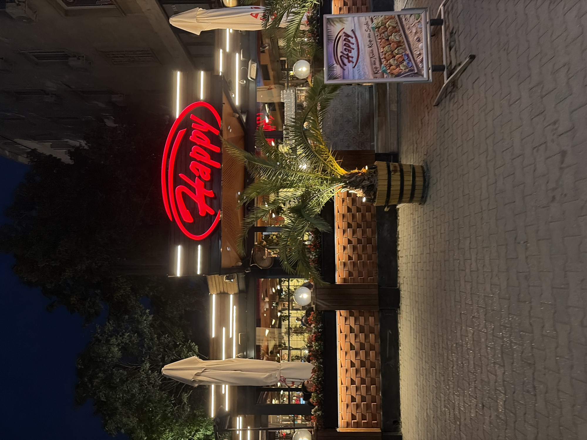 An exterior shot of a restaurant called "Happy" at night in Sofia, Bulgaria. The restaurant features a prominent red neon sign with the word "Happy" in a cursive font. The building facade is visible behind the sign, with windows and air conditioning units. In front of the restaurant, there are potted palm trees, planters with flowers, and outdoor seating areas with white umbrellas. A display board with images of sushi is placed on the sidewalk in front of the entrance. The ground is paved with interlocking bricks, and some areas appear wet, reflecting the lights. The overall ambiance is that of a dining establishment open for business in the evening.