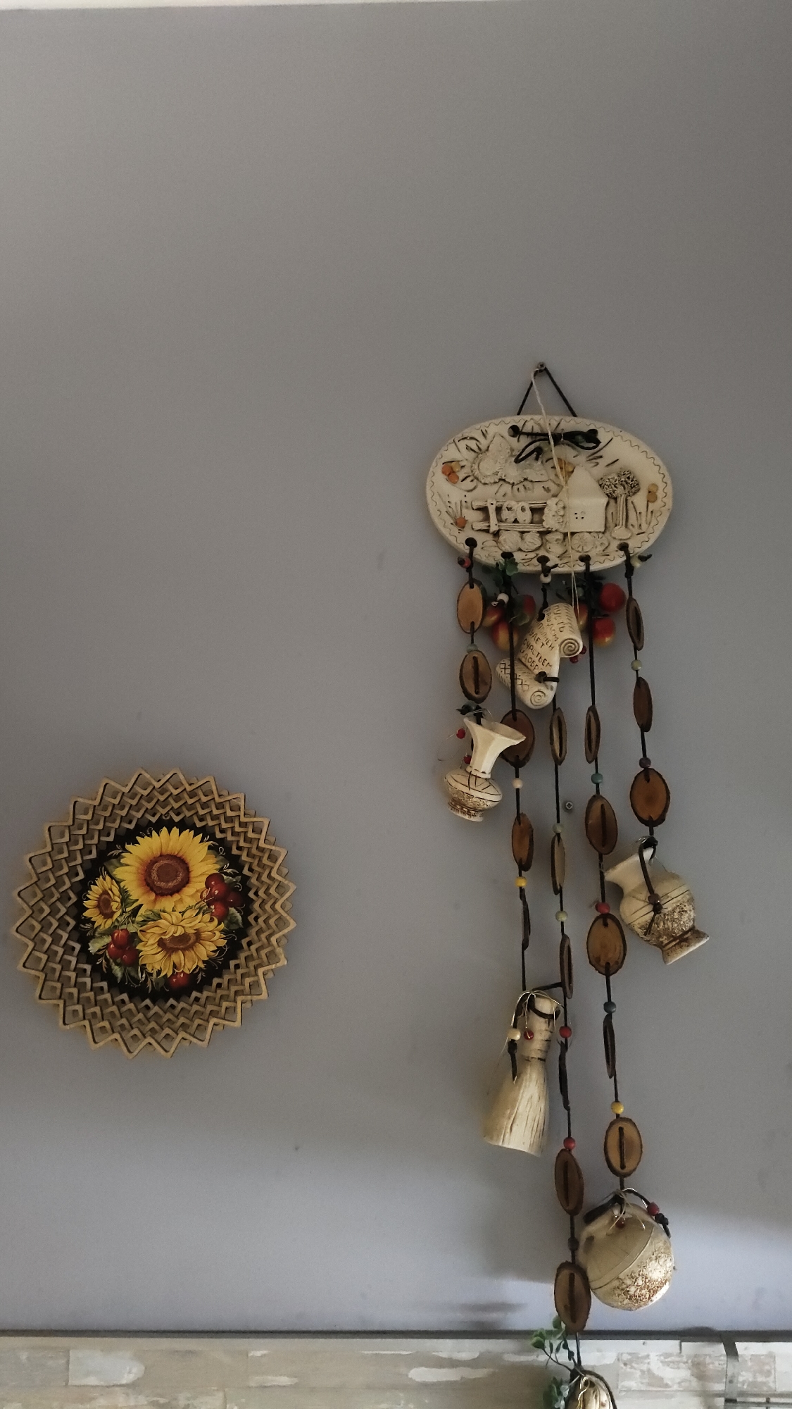 The image presents a section of an interior wall, likely in a residential setting. The wall is painted a neutral gray color. Two decorative items are mounted on the wall:
1. On the left, there is a circular wall plate featuring an arrangement of sunflowers and cherries. The plate has a dark background and a textured, geometric outer edge.
2. To the right of the sunflower plate, hangs a ceramic wall decoration. This piece is topped by an oval plaque with a rustic scene featuring a house and figures. Hanging below the plaque are strings of beads, wooden discs, and small, pottery-like shapes, resembling miniature vases or pitchers.
The lower portion of the image shows a section of countertop or a similar surface, possibly made of a light-colored, patterned material. A small green plant is also visible near the base of the wall decoration.
There is no indication of the location being Krasnoyarsk, Russia, in the image. The image provides no visible people, actions, or interactions.