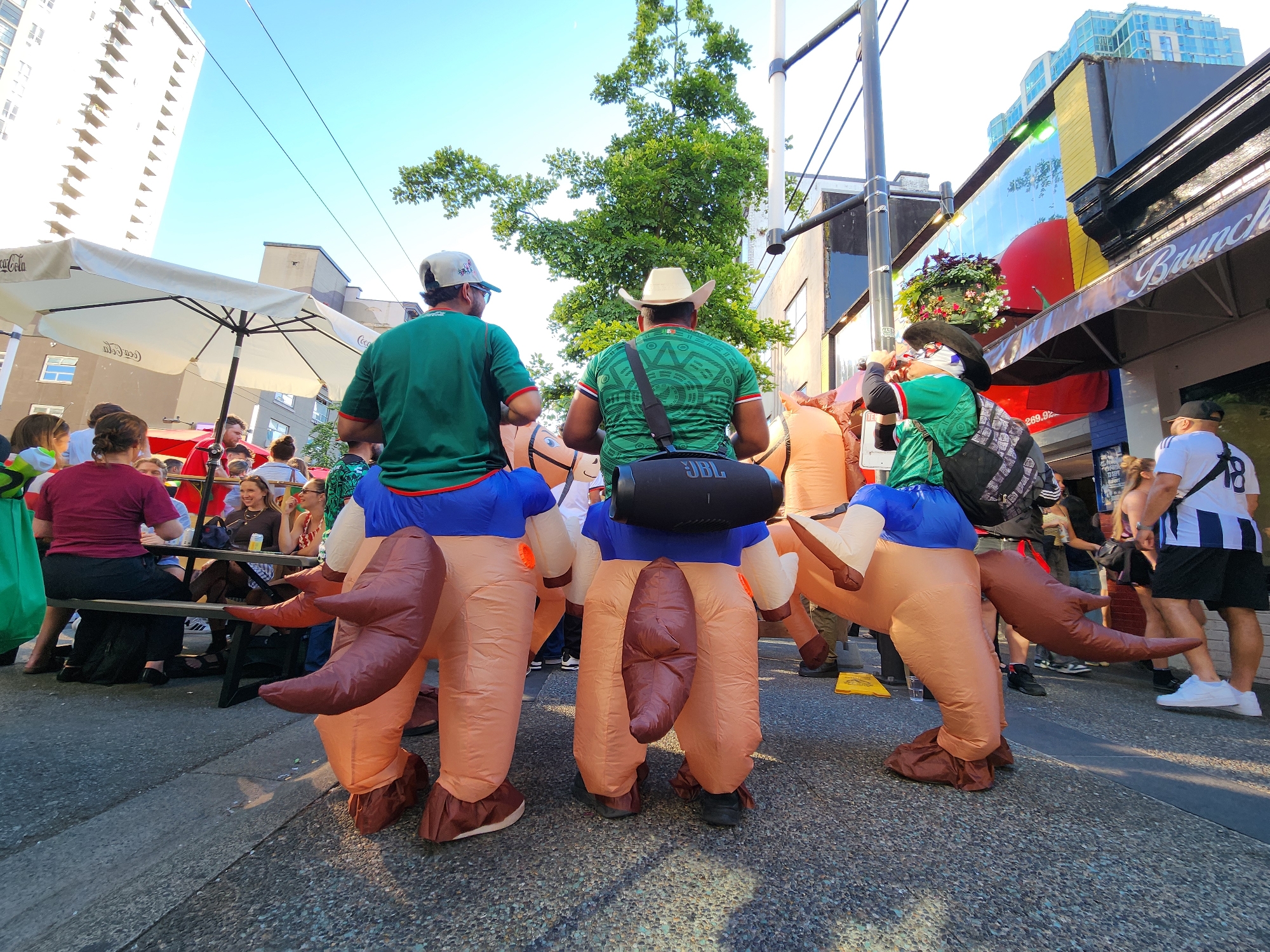 In a city setting under a clear blue sky, people are gathered on a paved street lined with buildings and trees. In the foreground, two individuals are wearing inflatable costumes that resemble horses from the hindquarters upwards. These costumes are tan and brown, with large, sculpted hind legs and tails. They are walking or standing on the street, facing away from the camera. One person in the center is wearing a green shirt with a patterned design and a straw cowboy hat, and has a black JBL speaker strapped to their chest. The other person to their left is wearing a green shirt and a baseball cap, also facing away. Behind them, other pedestrians are visible, some seated at tables under umbrellas. One building in the background has a vibrant mural or advertisement featuring a large red apple. The overall scene appears to be a public gathering or event in an urban environment.