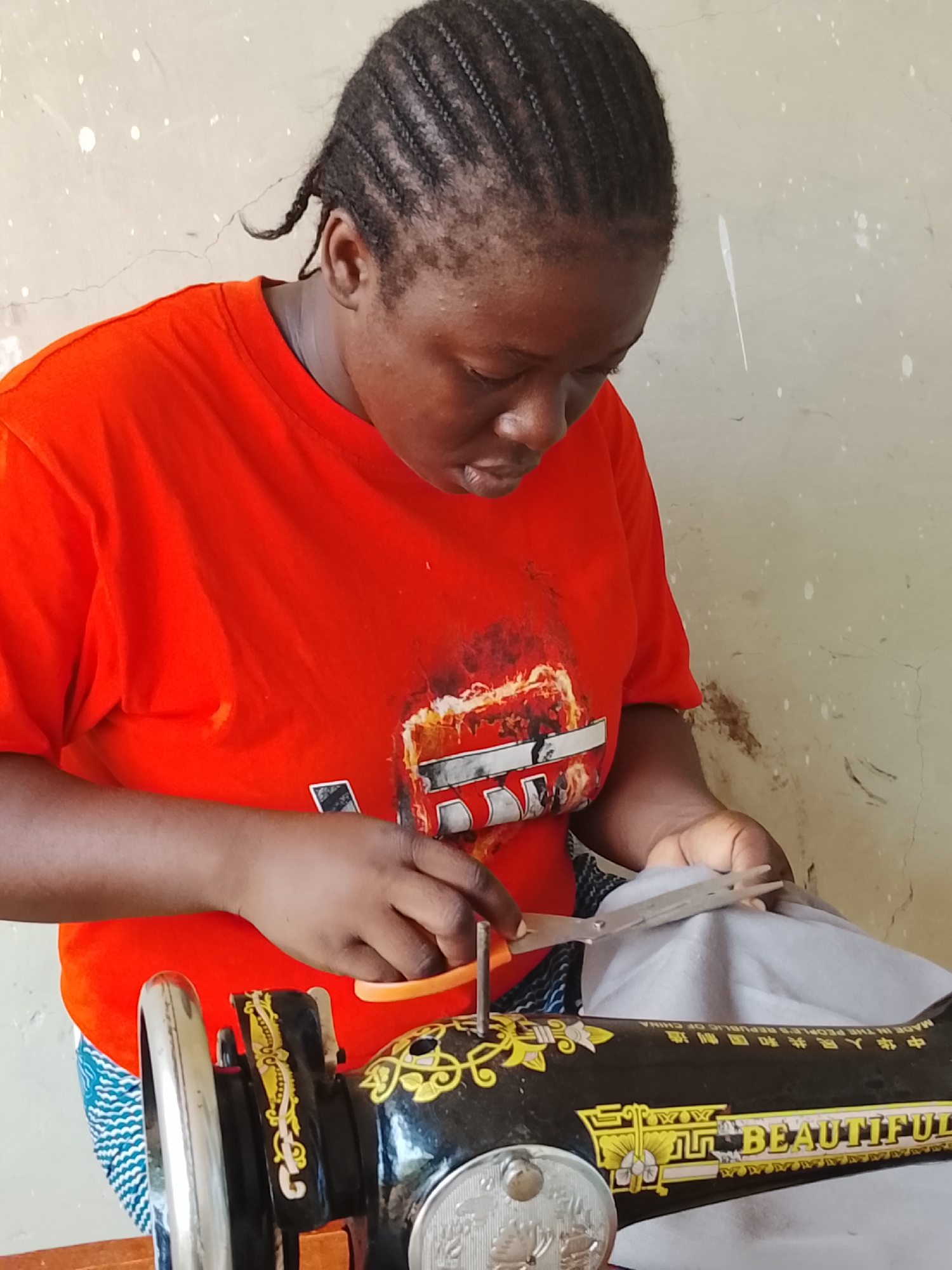 A woman with braided hair wearing an orange t-shirt is shown using scissors to cut gray fabric while standing at an old black sewing machine. The machine has the word "BEAUTIFUL" printed on it and what appear to be Chinese characters. The background is a plain beige wall. This image likely portrays a seamstress at work in Akwanga, Nigeria.