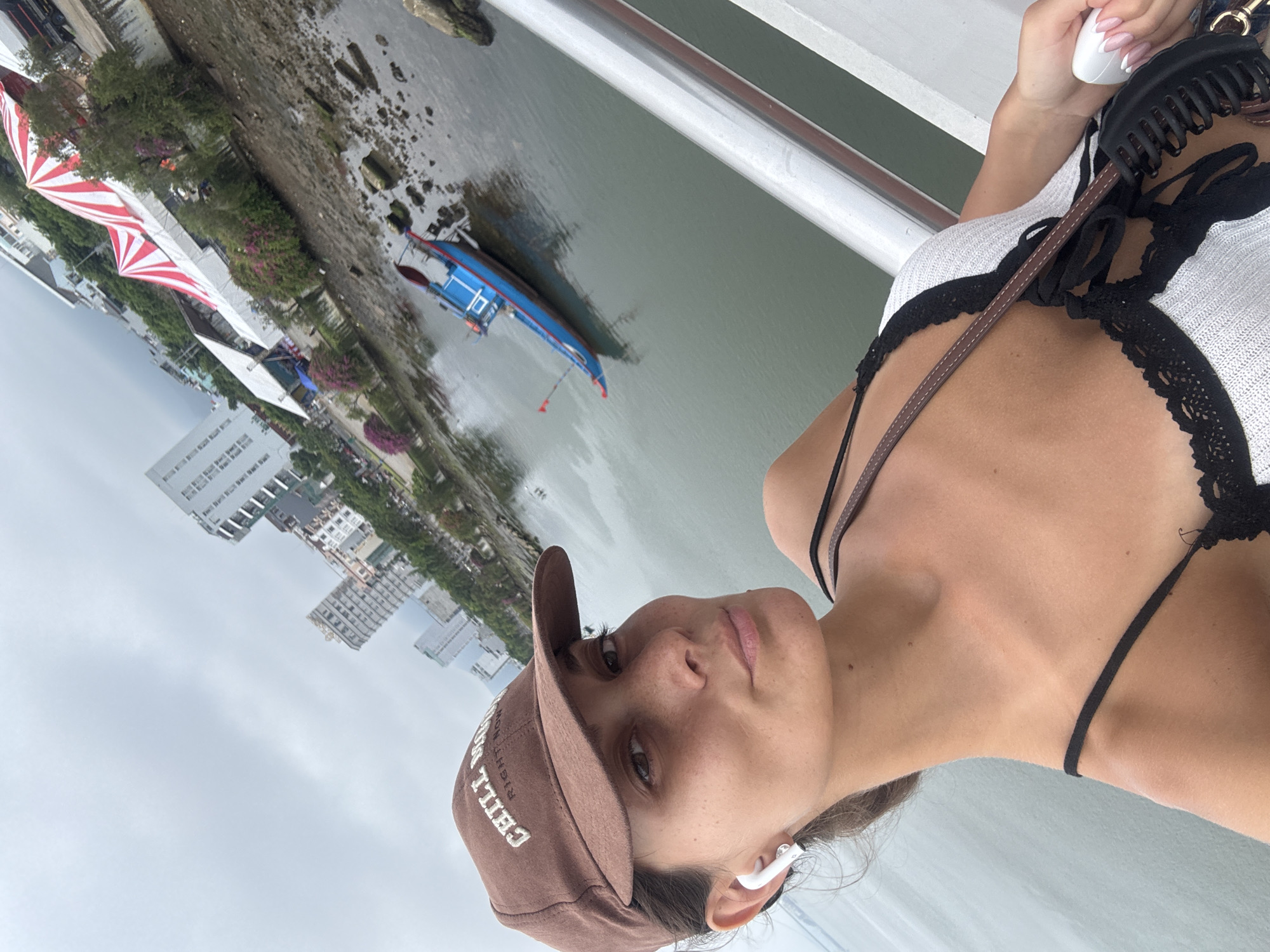 A person with light skin wearing a brown baseball cap and a white and black top is looking at the camera. They have light brown hair and are wearing an earbud in their right ear. A black purse hangs on their shoulder. The person appears to be on a bridge, as evidenced by the railing to their right. Below the bridge is a body of water with a blue boat and a shoreline covered in vegetation. In the background, buildings can be seen in the city of Đà Nẵng, Vietnam. There is also a section with red and white canopies. The sky is overcast.
