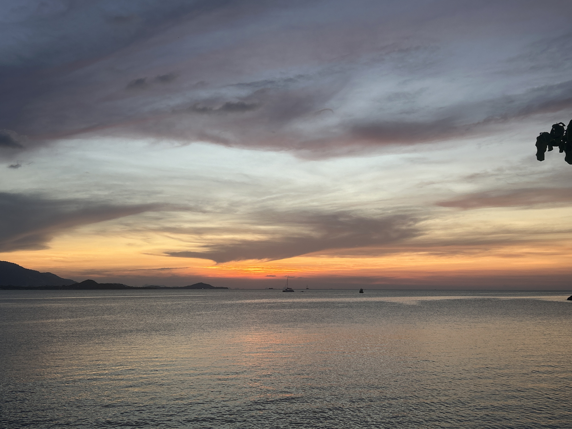 A tranquil seascape captures a sunset or sunrise over the waters near Baan Plai Laem, Thailand. The sky dominates the upper portion of the image, showcasing a dynamic interplay of clouds. The upper clouds are a mix of dark greys and purples, gradually lightening to soft greys and whites in the mid-sky. Towards the horizon, the sky erupts in vibrant oranges and yellows, hinting at the position of the unseen sun. Subtle pink hues also weave through the clouds above the brighter horizon.
Distant landmasses, appearing as dark, undulating silhouettes, stretch across the horizon, separating the glowing sky from the calm sea. The water reflects the muted light of the sky, appearing dark grey-blue with gentle ripples across its surface. Two boats are visible on the water: a prominent sailboat anchored or slowly moving towards the center-right, and another smaller vessel further to the right. These boats are silhouetted against the colorful horizon. A dark, out-of-focus mass, likely foliage or a tree branch, intrudes from the top right corner, suggesting the photograph was taken from under or near some natural cover. The overall scene evokes a sense of peace and natural beauty at dusk or dawn. No text is visible in the image.