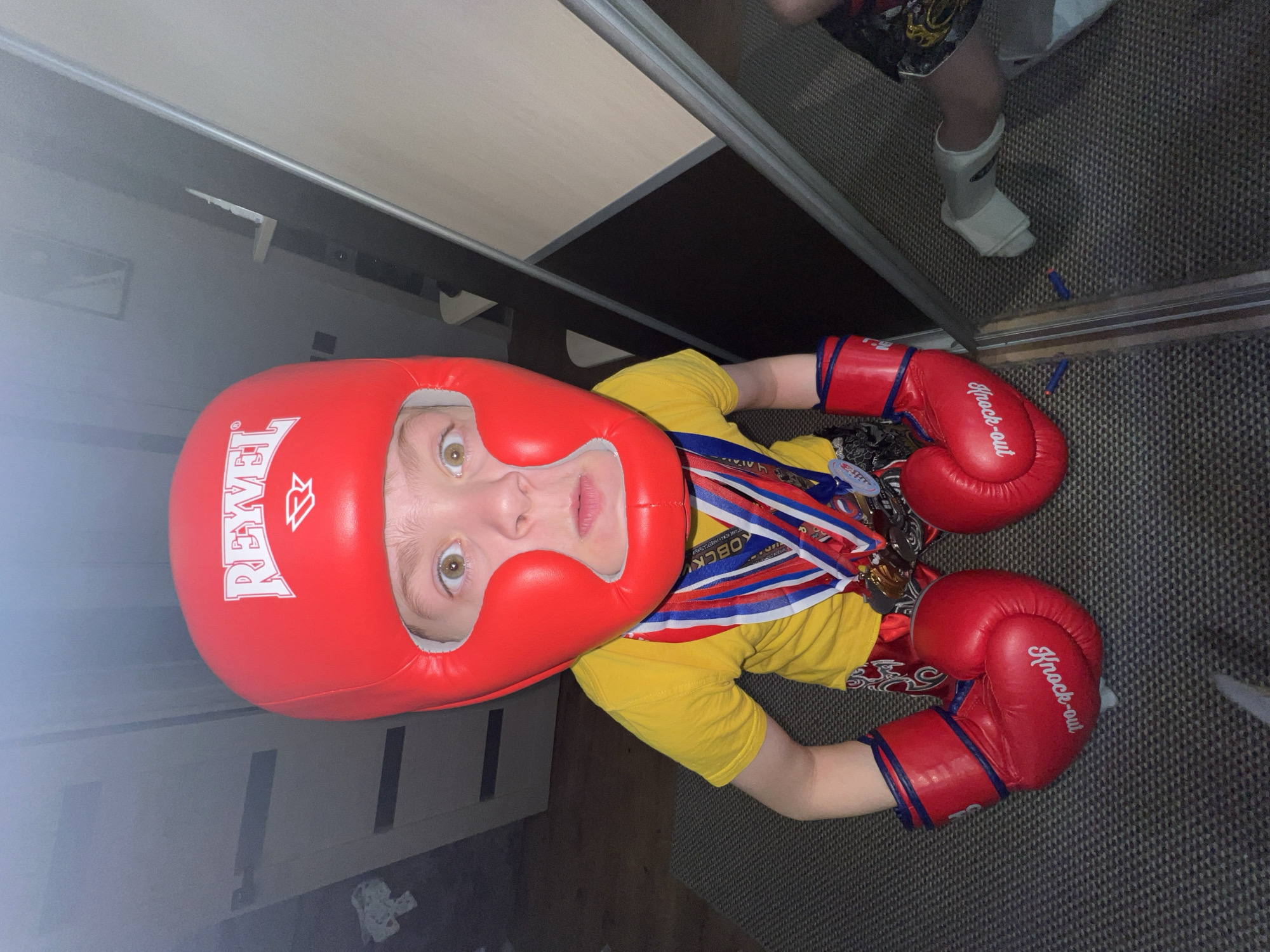 A young child wears a red boxing headgear and boxing gloves, along with a yellow shirt, red shorts with Asian lettering, and medals around their neck. The brand "REYMIV" is printed on the headgear, and the gloves say "Knock-out". Another person's legs are visible in the upper right corner, wearing black shorts and a white shin guard. The setting appears to be indoors, near a mirrored closet. Two blue darts are on the floor next to the mirror. The medal ribbons have the word "KOBOCK" on them, which is most likely "ВОЙСК" in Cyrillic, implying military sports or activities in Noviy, Russia.
