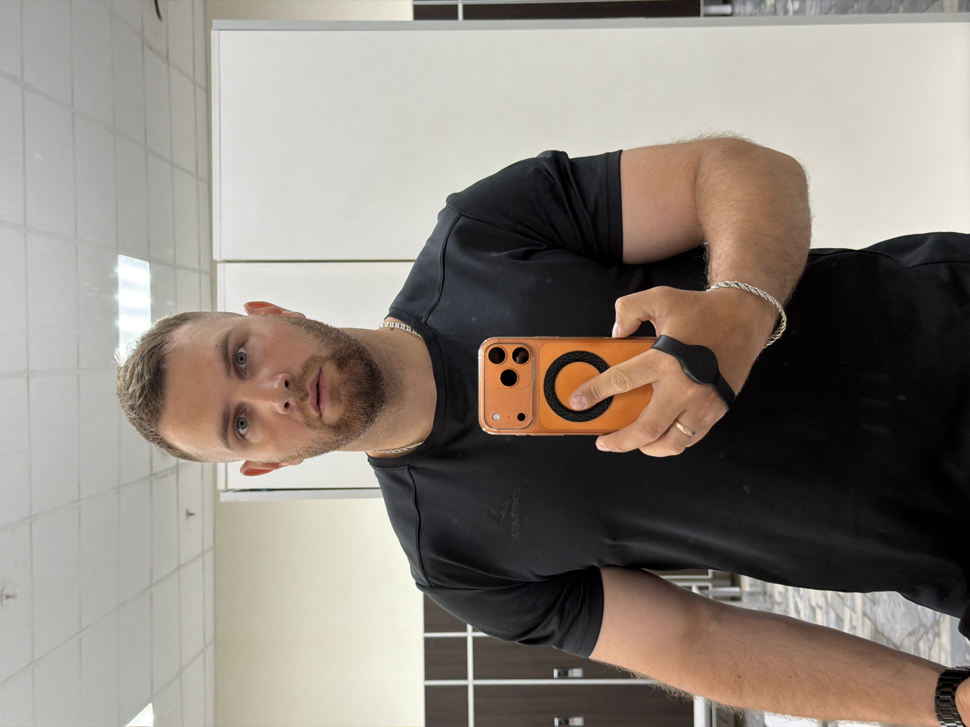A man takes a mirror selfie indoors, holding an orange smartphone. He is positioned slightly off-center to the left, looking directly into his phone's camera, which is reflecting his image. His expression is neutral with slightly parted lips.
The man has short, light brown hair and a neatly trimmed beard or stubble. He has light-colored eyes, possibly blue or grey. He is wearing a black short-sleeved t-shirt with a subtle, dark, almost indistinguishable logo on the left chest, which appears to be a stylized triangle and possibly letters like "XIMFC". Around his neck is a silver chain. His arms are visibly hairy. On his left wrist, he wears a braided silver bracelet, and on his right wrist, he has a dark-colored watch. A gold ring is visible on the ring finger of his left hand. The orange smartphone he holds has a black MagSafe-compatible ring on its back and features three camera lenses.
The background suggests an indoor public or commercial facility. To the upper left, there are white tiled walls, with some reflection from what might be overhead lights or windows. The upper right shows plain white panels or cabinets. In the lower background, there are dark brown or black lockers or cabinets, indicating a changing room, gym, or similar facility. The ceiling, partially visible in the reflection, features white square acoustic tiles with visible light fixtures.