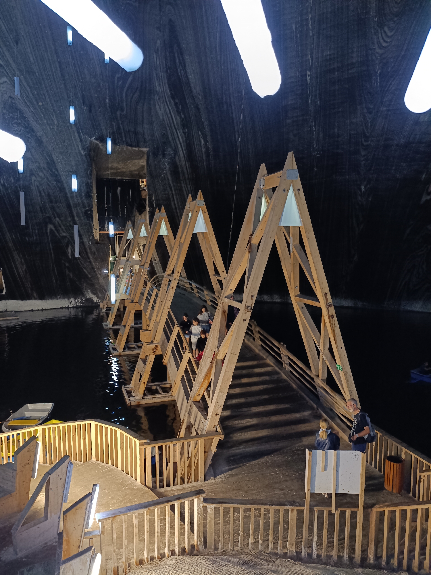 The image depicts an underground salt mine in Turda, Romania, transformed into a tourist attraction. The scene is dominated by the rough, striated walls of the salt cavern, illuminated by large, elongated light fixtures embedded in the ceiling and by smaller, rectangular lights hanging from wires. A dark, calm body of water fills the cavern floor, reflecting the lights and the wooden structure that spans it.
A prominent wooden bridge, constructed with a series of A-frame supports, crosses the water. The bridge is equipped with wooden railings and stairs, and several people are seen walking on it or standing at its edges. Some are looking out at the cavern, while others appear to be conversing. In the foreground, a wooden walkway with railings leads down to the water's edge, with several benches also visible. A small, white rowboat is moored near the walkway.
The lighting suggests it is likely nighttime or the cavern is artificially lit, as there is no natural daylight visible. The atmosphere appears serene and perhaps a bit awe-inspiring, given the unusual and grand setting. The overall impression is of a unique tourist destination that harmoniously blends natural geological formations with man-made structures and recreational elements. No discernible text is visible in the image.