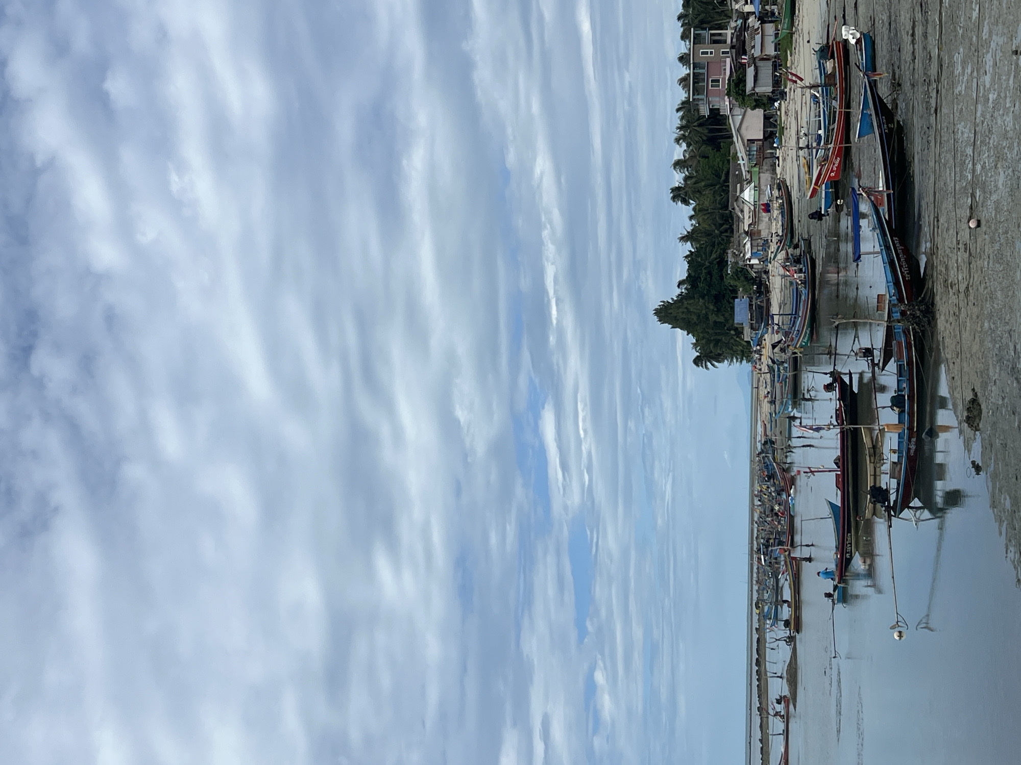 The image depicts a coastal scene, possibly in Baan Hua Thanon, Thailand, given the presence of traditional longtail boats. The foreground displays numerous beached boats on a mudflat or sandy shore during low tide. A line of buildings and trees is visible along the shoreline behind the boats. The sky is heavily overcast with striated clouds. The water reflects the sky, creating a muted color palette. There are no clearly visible people in the scene.