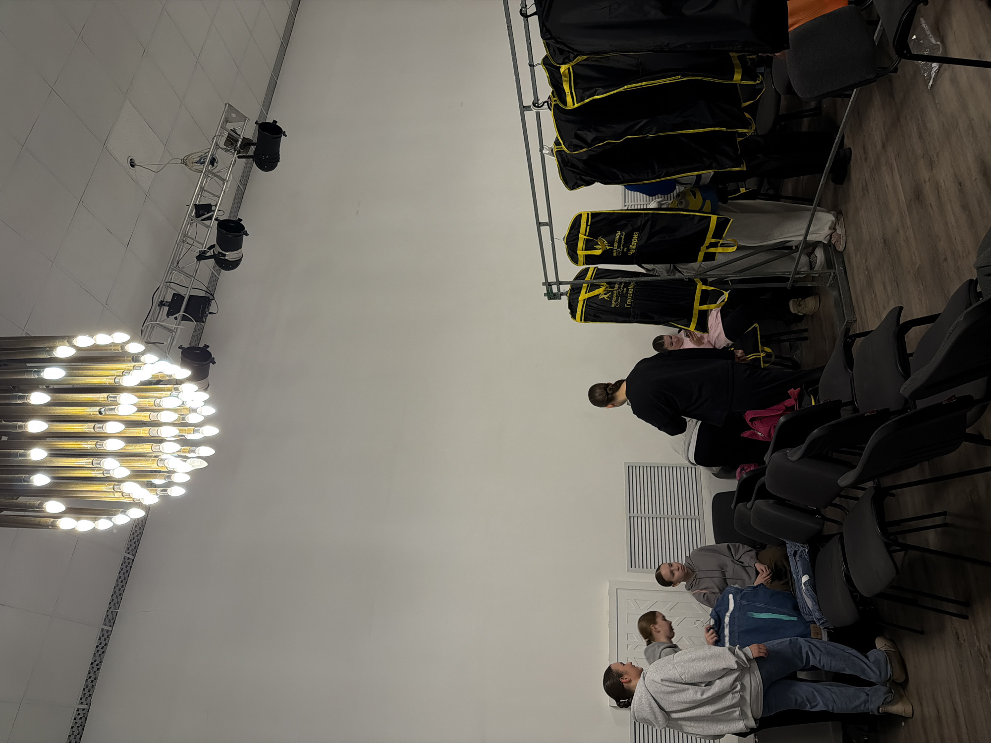 The image depicts the interior of a room in Perm, Russia.
A large chandelier illuminates one side of the room. Stage lighting is present. Clothes racks holding black and yellow garments are visible. Several people are present, including a woman in black and a group of three seated individuals. There are rows of black chairs in the room. A white door is visible in the background.