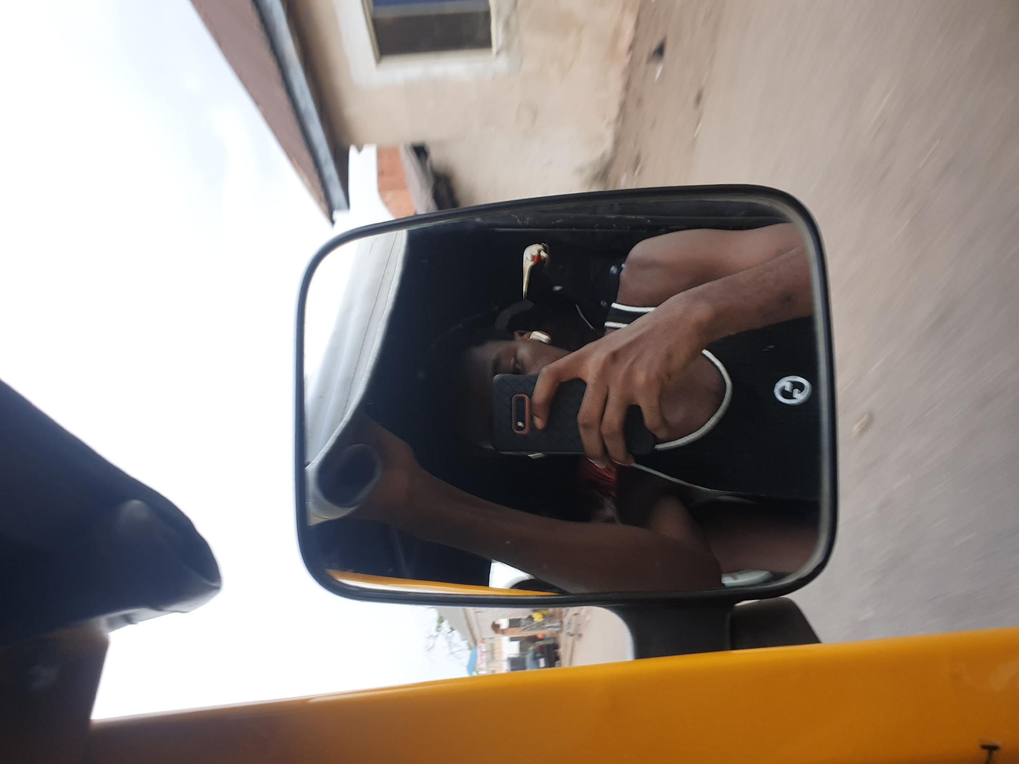 The image captures a selfie taken from the reflection in a rearview mirror, likely from a moving tricycle (Keke Napep) in Lokoja, Nigeria. The central subject is the black-framed rearview mirror, reflecting the individual taking the photo.
Within the mirror's reflection, a person is visible, wearing a dark sleeveless top that features a white circular logo containing a stylized symbol, possibly an 'S'. A white earbud is seen in their right ear. Their face is partially obscured by a black smartphone held in their hands, which has a textured case and a camera lens with an orange/red accent. The person's bare arms are visible.
Outside the mirror, the upper left portion of the image shows a bright, overcast sky. On the right, parts of buildings with light-colored walls, windows, and sections of brick, along with a brown roof overhang, suggest an urban or roadside setting. A yellow bar, likely a handle or part of the vehicle's frame, is prominent in the lower part of the image, with additional dark gray vehicle components. Motion blur on the background elements, particularly the ground and buildings seen outside the mirror, indicates that the vehicle is in motion. The scene appears to be set during the daytime under bright conditions.