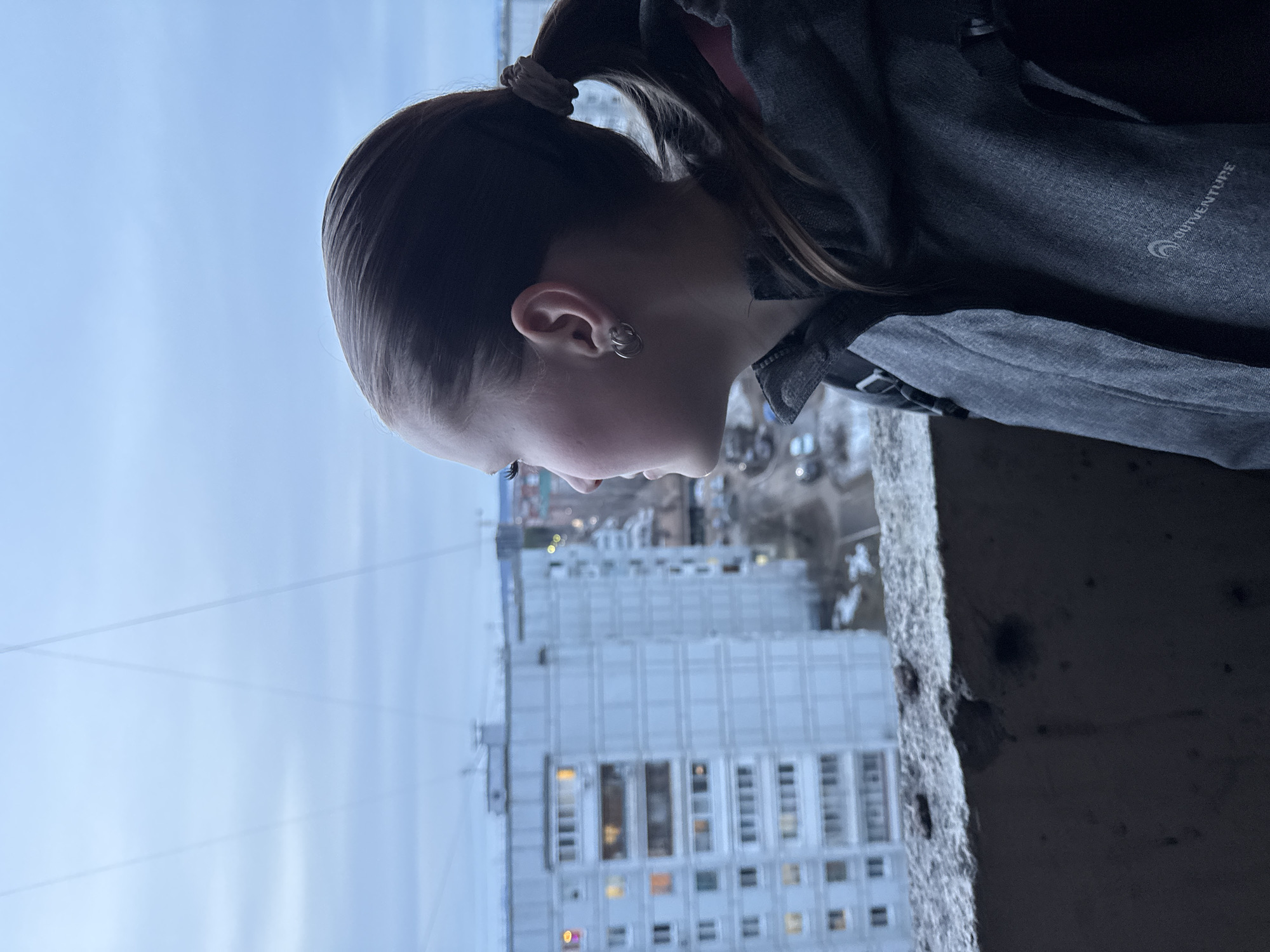 A young person is looking out from a high vantage point, possibly a balcony, in the city of Chaikovsky, Russia. The person, with their hair pulled back into a ponytail, is wearing a grey jacket marked with the word "OUTVENTURE." Below, a multi-story apartment building is visible, indicating an urban residential area. The sky is overcast, and the overall scene suggests a cool or cold climate.