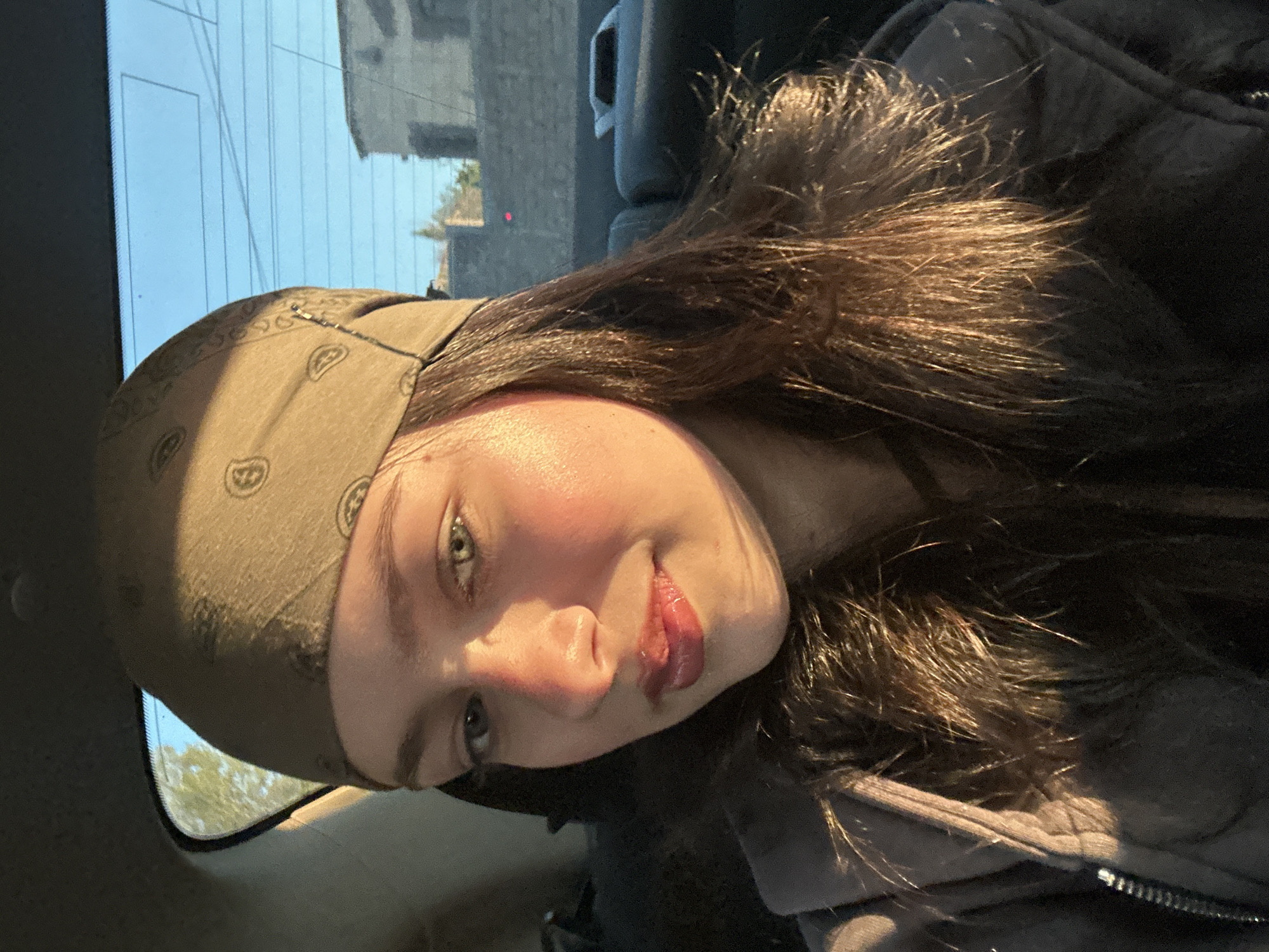 The image is a selfie of a young woman wearing a light brown bandana with a repeated pattern. She has brown hair, and her face is illuminated by sunlight. She is wearing dark clothing with a lighter colored jacket partially visible. The background appears to be the inside of a car with a window showing a building and blue sky. The specific location in Yerevan, Armenia, cannot be determined from this image.