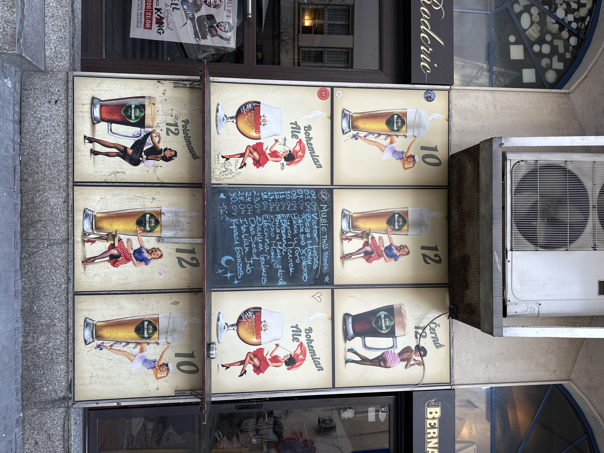The image shows a vertical shot of a building's exterior in Sofia, Bulgaria. Nine Bernard beer advertising posters are prominently displayed, each featuring a different pin-up girl posing with a glass of Bernard beer.
Adjacent to the posters is a chalkboard with a list of musical performances scheduled for the month. An air conditioning unit is mounted next to the posters on the wall. The image also captures a sign reading "Rodcric," parts of posters indicating "Jazz Kang", "CERT," and "Bulgaria". A storefront with a Bernard Beer sign and window displays below the advertisement posters is visible, as well as the building's exterior details and stonework.