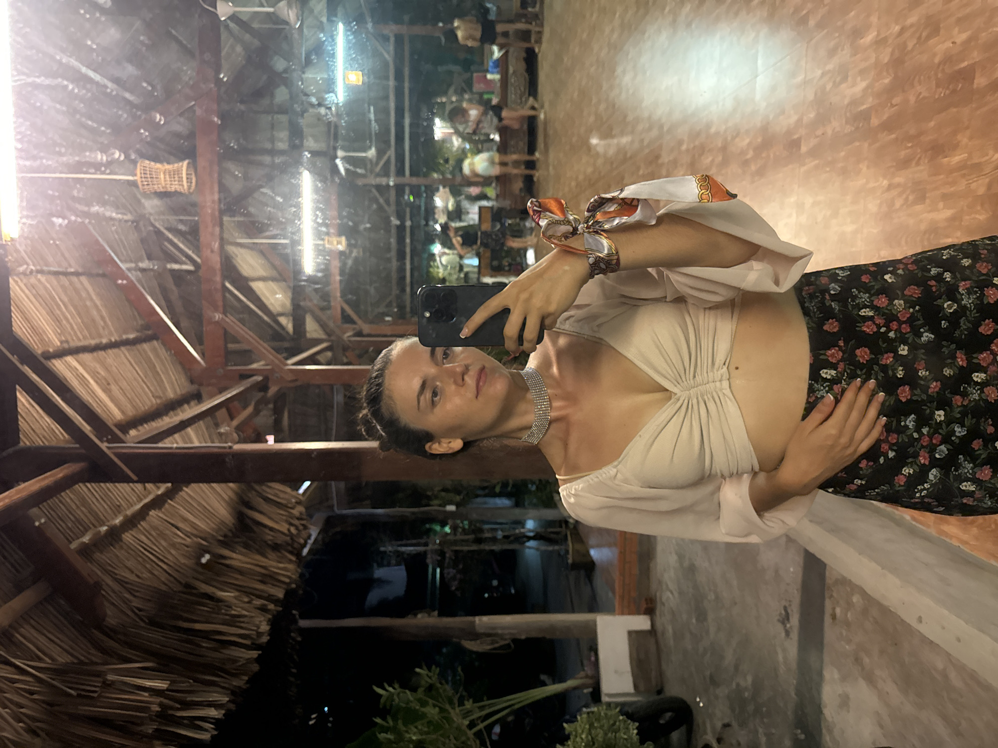 The image shows a woman taking a selfie in what appears to be a restaurant in Phu Thuy Ward, Vietnam. She is holding a smartphone up to her face. The woman is wearing a light-colored top and a floral print skirt. She also has a necklace. In the background is a dining area with a thatch roof. People are seated at tables and chairs. The ceiling has wood beams.
