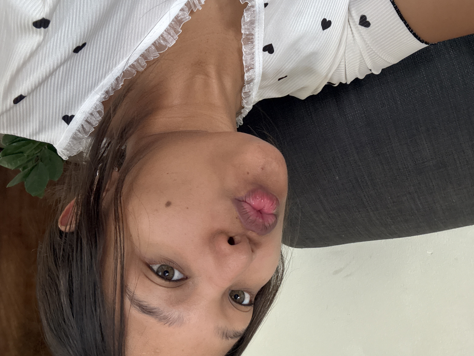 A close-up, upside-down selfie shows a woman with fair skin, dark brown hair, and brown eyes puckering her lips for a kiss. She has a small mole on her left cheek. She is wearing a white top with small black heart patterns and a delicate white lace trim around the neckline. Part of a dark gray or black cushion or surface is visible in the upper right corner. There are green leaves visible near the left side of her head. The background behind her is a light off-white color.
