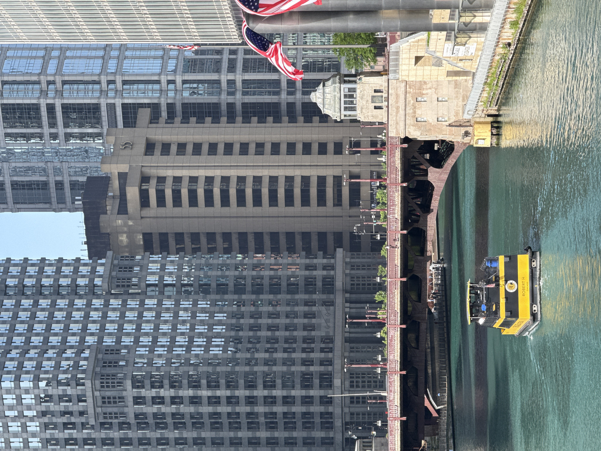 This is a medium shot of the Chicago River in Chicago, Illinois, USA, on a clear day. In the foreground, a yellow and black tour boat named "ROBERT H" is traveling away from the viewer, leaving a wake in the turquoise water. The boat has the name "CHICAGO" printed on its stern. Behind the boat, a red truss bridge spans the river, with its arches visible. A small, octagonal guardhouse with a pointed roof sits on the south bank of the river, adjacent to the bridge abutment. Tall modern buildings with reflective glass facades dominate the background. American flags are visible on poles attached to cylindrical structures on the right side of the image. The overall scene depicts a typical urban riverfront environment in Chicago.