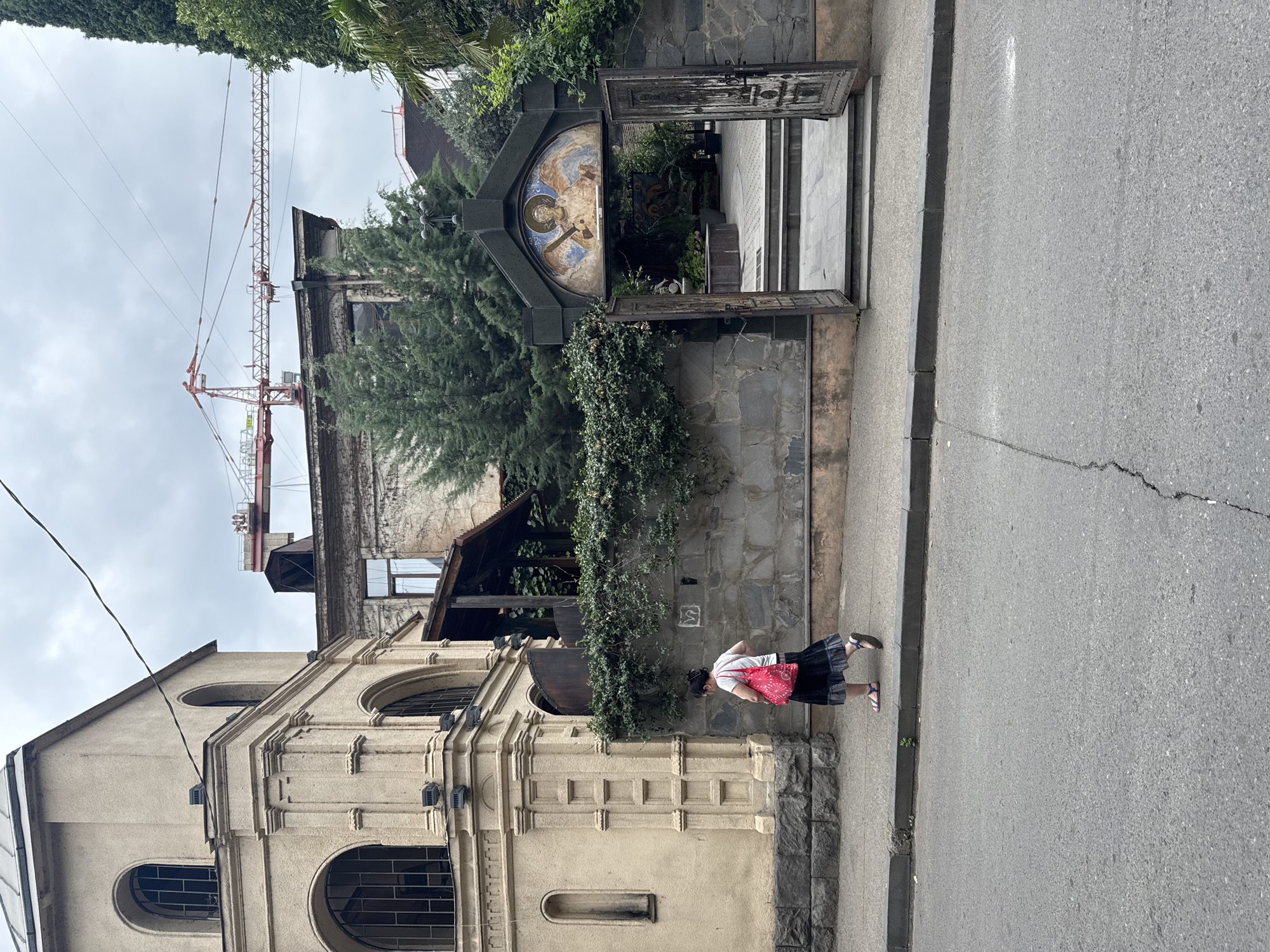 An overcast day in Tbilisi, Georgia, captured from a street level perspective. In the foreground, a cracked asphalt road runs horizontally across the frame. To the left, a woman with dark hair pulled back walks away from the camera, wearing a white t-shirt, a dark patterned skirt, blue sandals, and carrying a bright pink tote bag. She is walking on a sidewalk beside a light-colored building with arched windows and decorative molding. Adjacent to this building, a stone wall topped with dense green foliage separates the sidewalk from an entryway. This entryway features an arched portal adorned with a religious fresco of an angel, and flanked by two weathered, open metal doors. Beyond the doors, a paved courtyard with stairs is visible, along with lush greenery. In the background, parts of older buildings are visible, one covered in ivy, and a large red construction crane stands prominently against the sky, indicating ongoing development in the city.