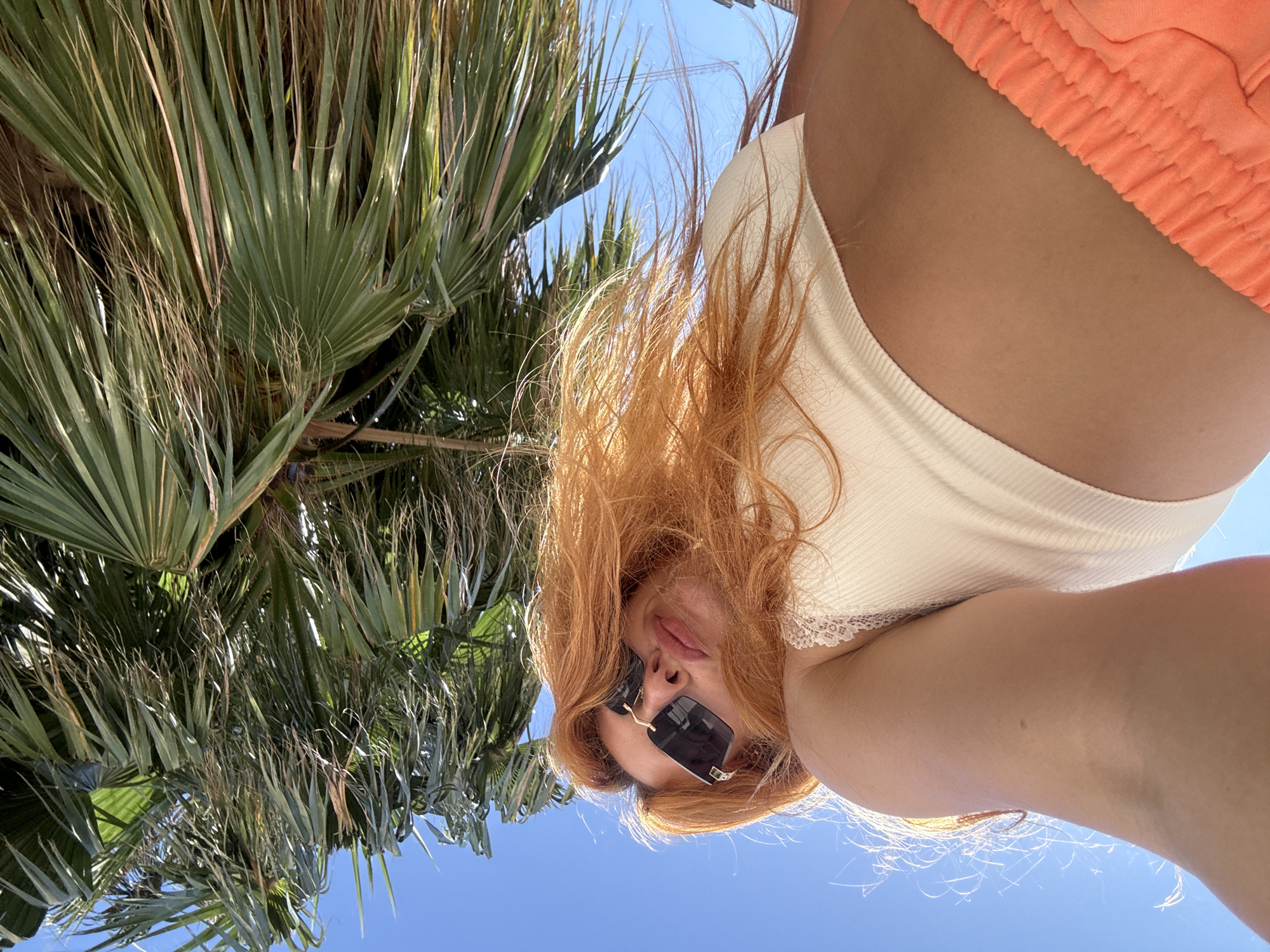 The image displays a woman captured in an outdoor, low-angle selfie under a clear blue sky in Budva, Montenegro. She possesses long, wavy, reddish-blonde hair, illuminated by bright sunlight. The woman wears dark, rectangular sunglasses with gold-colored accents on the temples, and her lips are slightly parted. She is dressed in a light-colored, possibly cream or off-white, ribbed crop top featuring white lace trim on the upper edge. The lower right portion of the image reveals the ruched, elasticated waistband of bright orange shorts. Above the woman, the upper half of the frame is dominated by the dense, green fronds of a palm tree, some exhibiting dried, straw-like elements, contrasting with the vibrant blue sky. The perspective is directed upwards, presenting the woman and the overhead foliage.