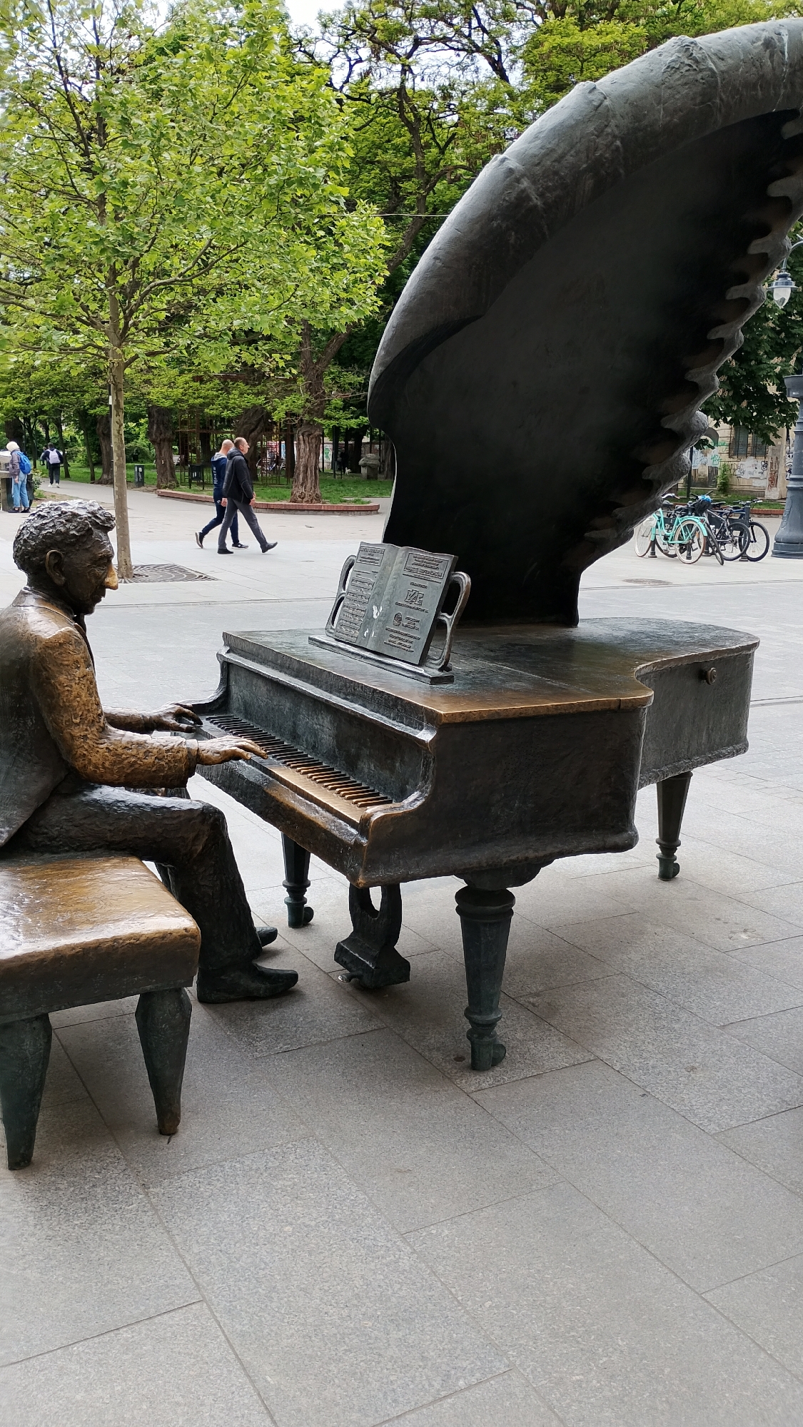 The image captures a life-sized bronze sculpture of Arthur Rubinstein playing a grand piano, located on Piotrkowska Street in Łódź, Poland. The statue depicts Rubinstein seated on a bench in front of the piano, with his hands positioned as if playing the keys. The piano has an open lid and a music stand holding sheet music. In the background, there are trees, two people walking, and bicycles parked near a lamppost. The ground is paved with grey stone tiles.