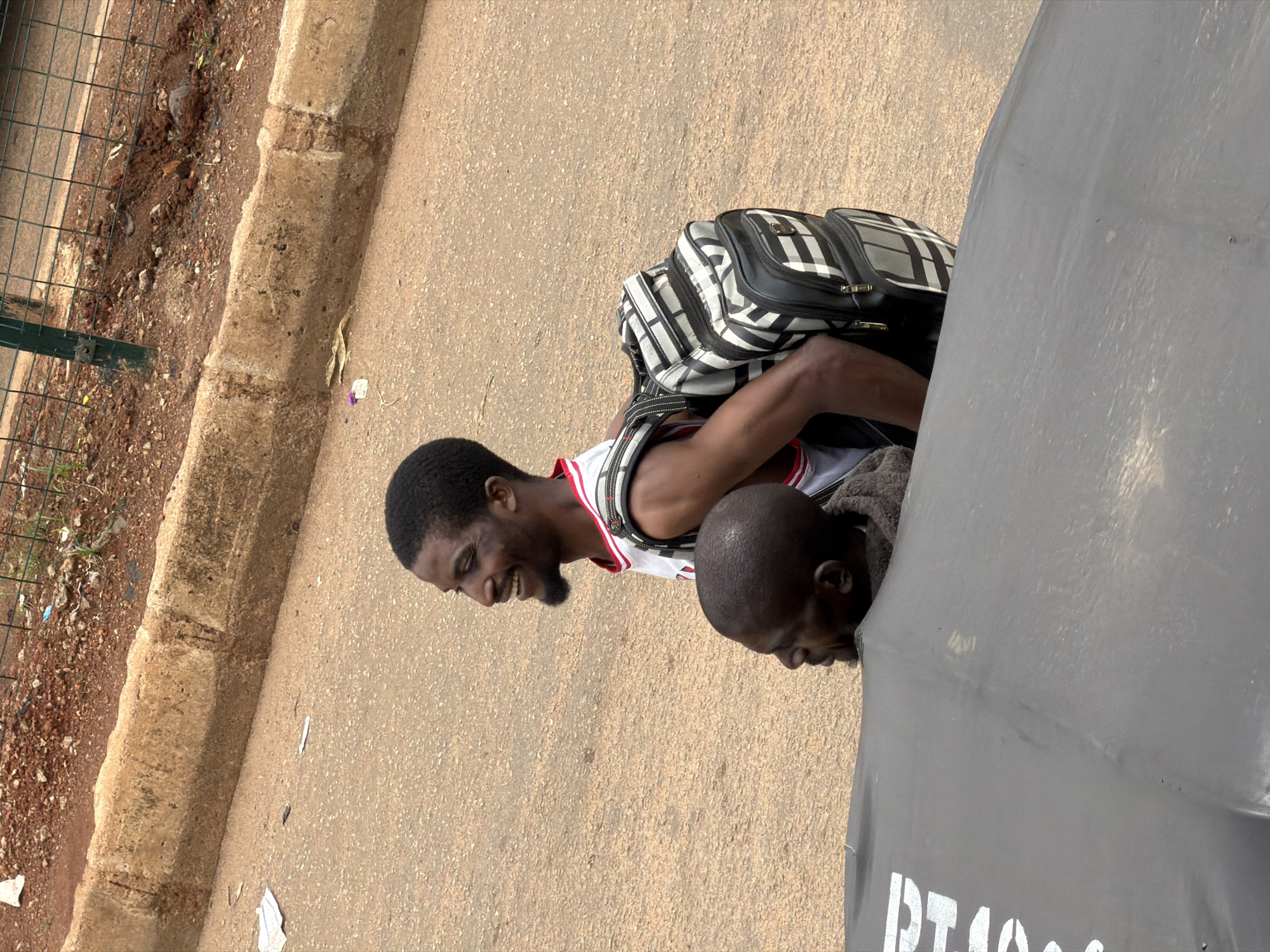 Two men are on a street in Bukuru, Nigeria. The man in the foreground is wearing a white tank top with red trim and has a beard. He is smiling and looking down. He is carrying a black and white checkered backpack. Behind him, another man with a shaved head is partially visible, also looking down. They appear to be near a large, dark object, possibly a vehicle or a large umbrella. The ground is paved, and there is a concrete curb to the left, with loose dirt and debris next to it. A metal fence is also visible to the left. The lighting suggests it is daytime. There is some white text visible on the dark object, which appears to be "PT1000" stenciled in white.