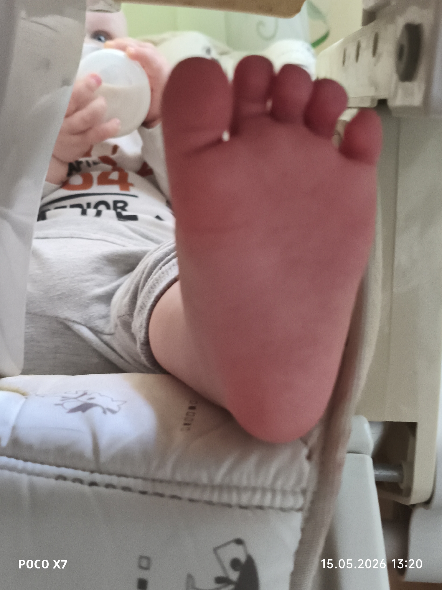 A baby sits in a high chair holding a bottle. The baby's foot is prominently displayed towards the viewer. The high chair has a patterned seat cushion. The image was captured on May 15, 2026, at 13:20 using a POCO X7. The location is not specified, and therefore cannot be verified to be in Chaikovsky, Russia.