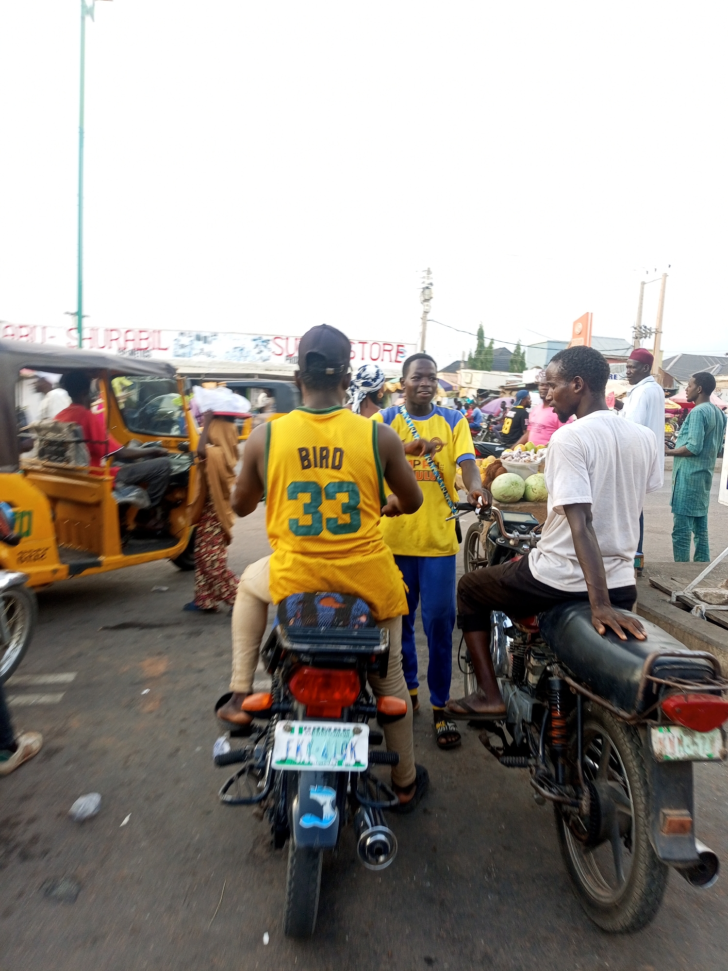 The image depicts a street scene in Tempure, Nigeria. A yellow three-wheeled vehicle is to the left of the frame. A man in a yellow jersey labeled "BIRD 33" and khaki pants sits on a parked motorcycle, facing away from the viewer. Next to him is a man wearing a yellow and blue outfit, standing and looking at a man in a white shirt and black pants who is sitting on another motorcycle. In the background, several other people are visible, along with a fruit stand displaying watermelons and a building with the words "STORE" visible on its facade. The motorcycle in the foreground has a license plate, IKY-419K.