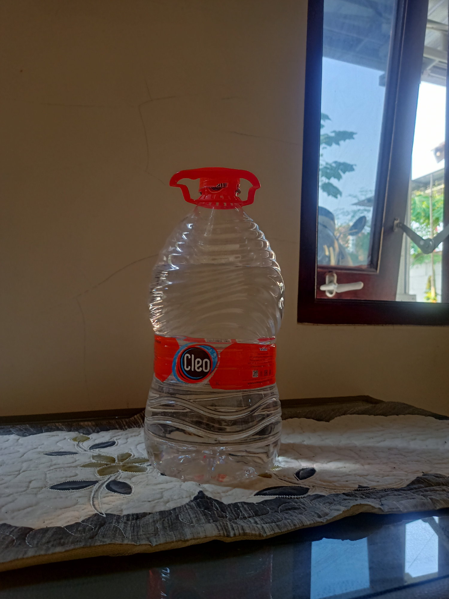 A half-filled, clear plastic bottle of Cleo brand mineral water stands upright on a table. The bottle has a red cap and a red and blue label. The table is covered with a quilted fabric placemat featuring a floral embroidery in shades of green and gray. Behind the bottle, a plain, light brown wall is visible, with subtle cracks and lines. To the right of the bottle is a window with a dark brown frame. The window is partially open, revealing a blurred outdoor scene with trees and a bright sky. A white window latch is visible on the frame. The overall lighting suggests an indoor setting with natural light coming from the window. There are no people or animals present in the scene.