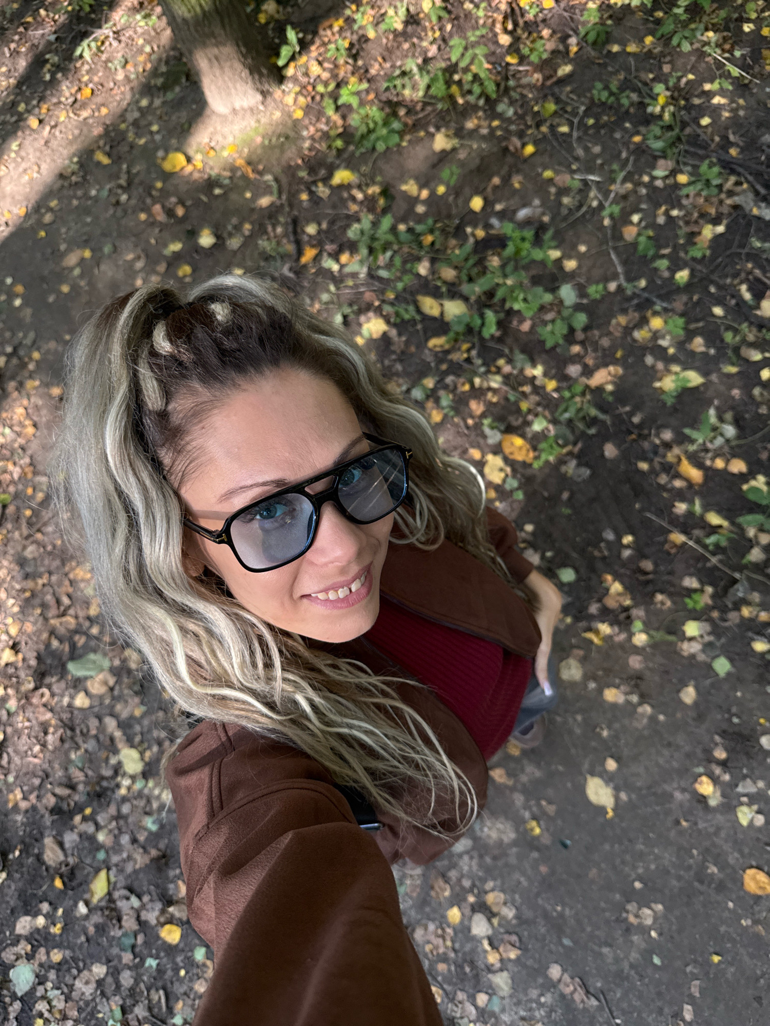 A woman with blonde, wavy hair styled in a high ponytail, wearing large, dark-rimmed glasses and a brown jacket over a maroon top, is pictured from a high-angle, looking up at the camera with a smile. She is standing outdoors on a path covered with fallen leaves, suggesting an autumn setting. The ground is dark and uneven, with scattered green foliage and yellow leaves. A tree trunk is visible in the upper left corner of the frame, with dappled sunlight falling on the path. The image appears to be taken in a park or wooded area within the city of Dumino, Russia.