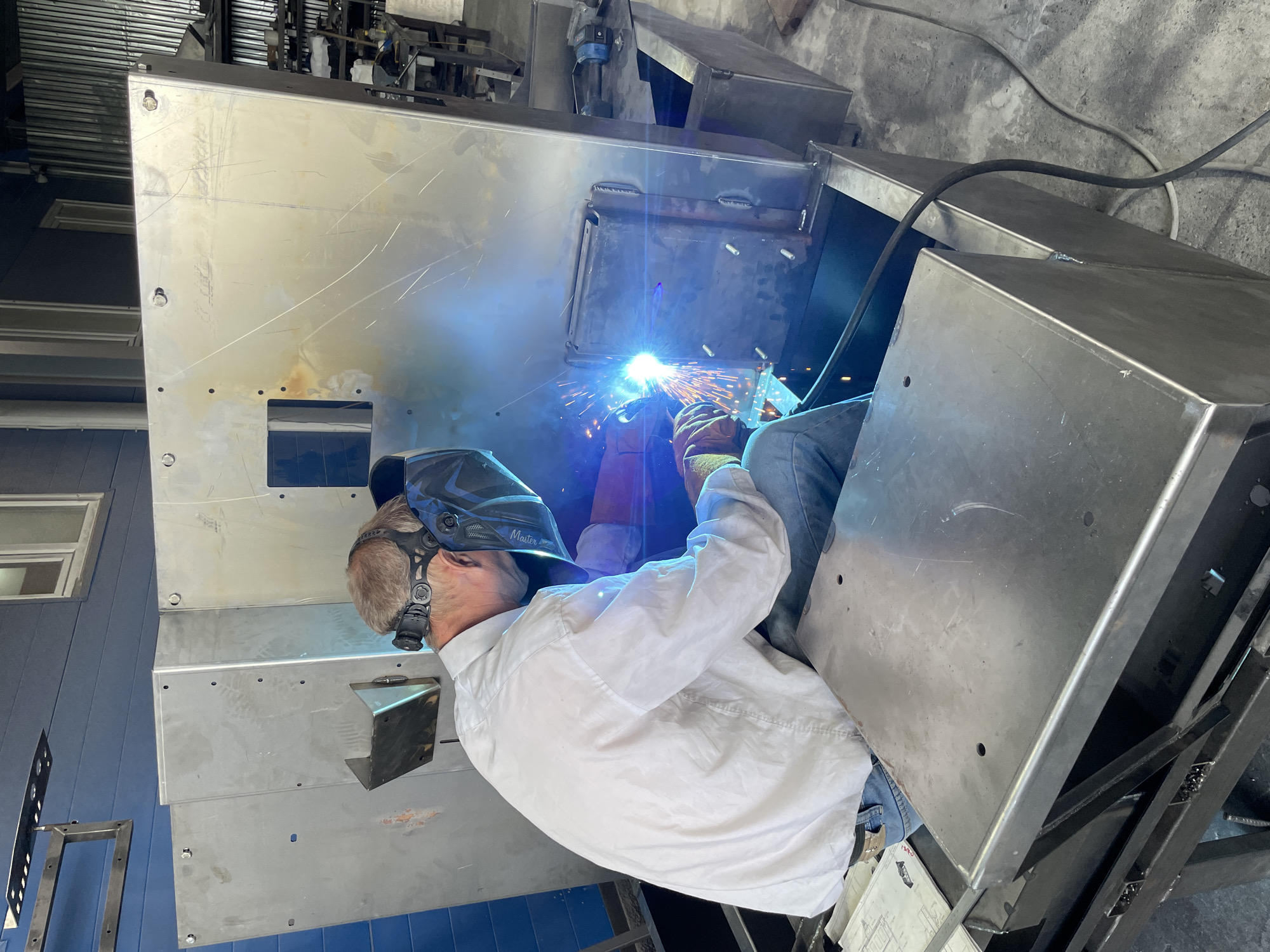 A person wearing a white long-sleeved shirt, blue jeans, welding mask, and gloves is welding a large, unfinished metal structure. The welding process is creating a bright blue arc and orange sparks. The structure appears to be a large, box-like metal cabinet or enclosure, possibly part of a larger machine. The environment appears to be a workshop or industrial setting, with concrete flooring and other metal structures visible in the background. The lighting is bright, likely from overhead industrial lights and the welding arc. The image is taken from a slightly elevated, rear-view perspective of the person welding. The location is not specified as Сокуры, Russia.