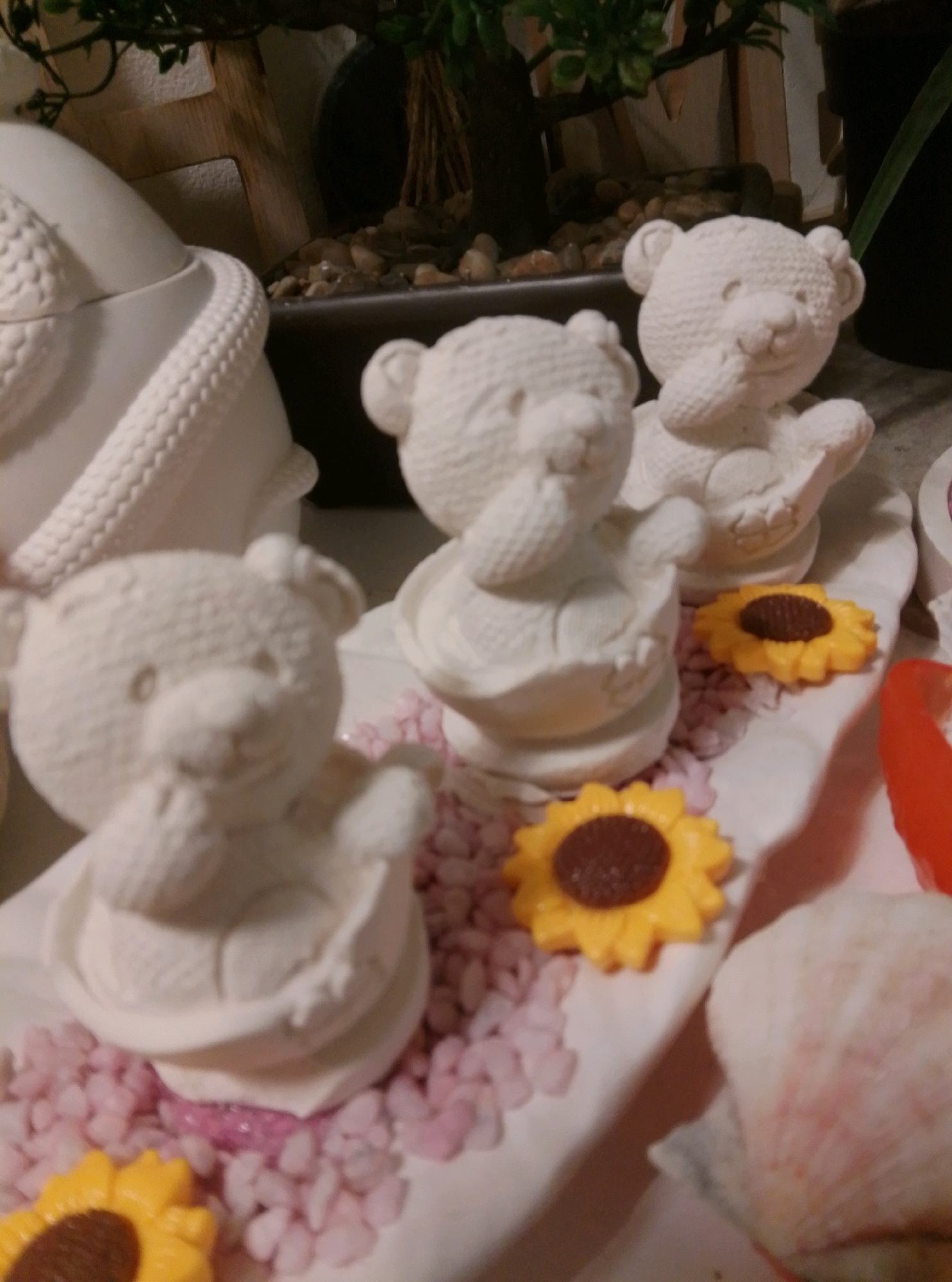 The image depicts a close-up view of a decorative arrangement featuring several white ceramic teddy bear figurines. The bears are positioned on a white surface, possibly a tray or platter, which is adorned with small pink decorative stones. Two yellow sunflower-shaped decorations are also visible, one in the foreground and another slightly further back. In the background, a potted plant with green foliage is visible, along with some small stones and what appears to be a wooden structure. The overall scene suggests an indoor setting, possibly a home or a craft store. There are no individuals present in the image. The lighting is somewhat dim, and the focus is on the teddy bear figurines and their immediate surroundings. The image is devoid of any information that would allow for its localization in Wedel, Germany, or any other specific city.