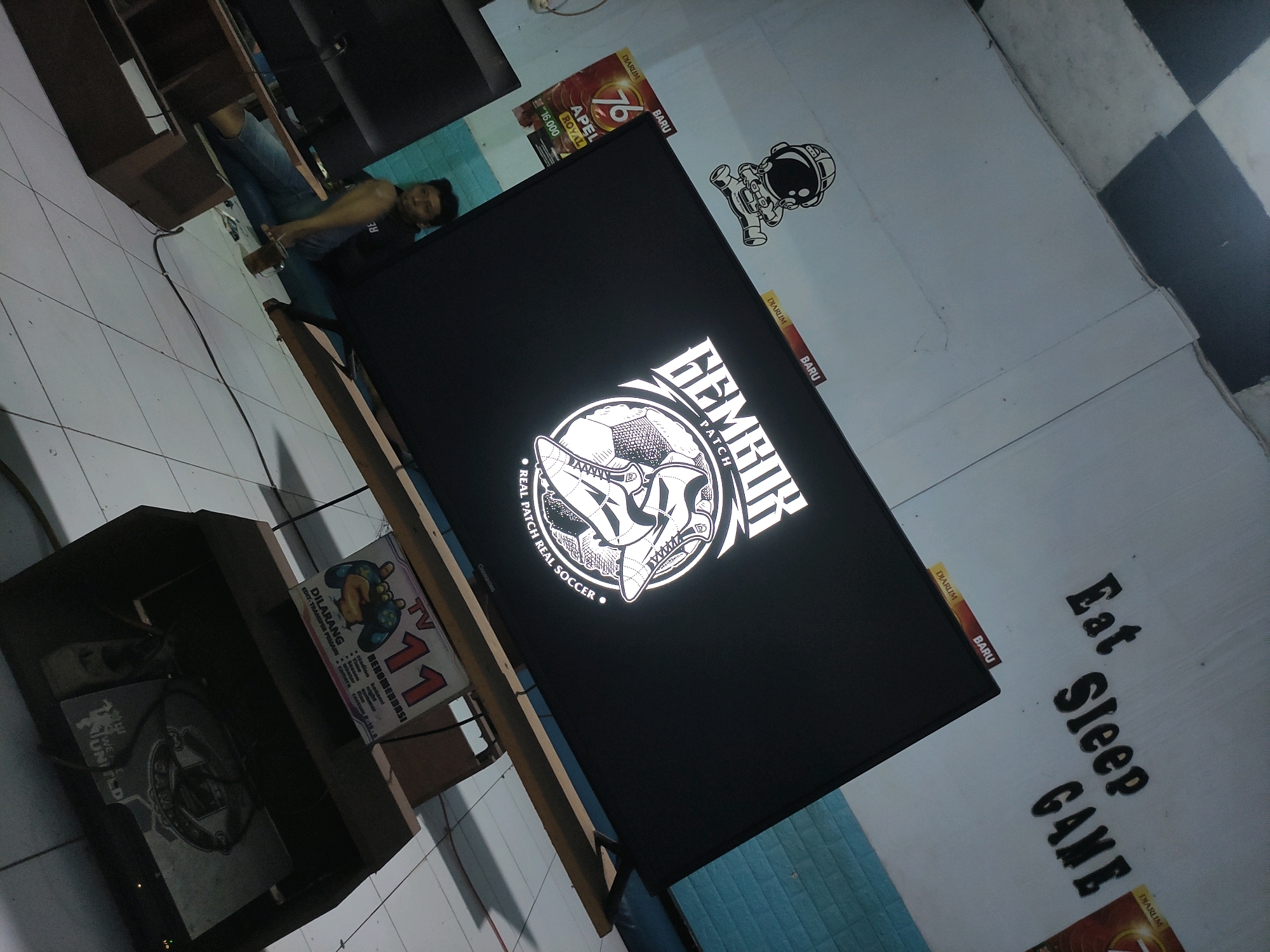 In Ngrendeng, Indonesia, a medium shot captures a person reclining on the floor with a glass in their hand. They are wearing jeans and a dark t-shirt with text on it. Beside them is a large black television screen displaying a white logo with text. In the background, a shelf unit is visible, along with various promotional materials and a black and white checkered tiled floor. To the right of the television, the words "Eat Sleep GAME" are written on the wall in large, dark lettering.