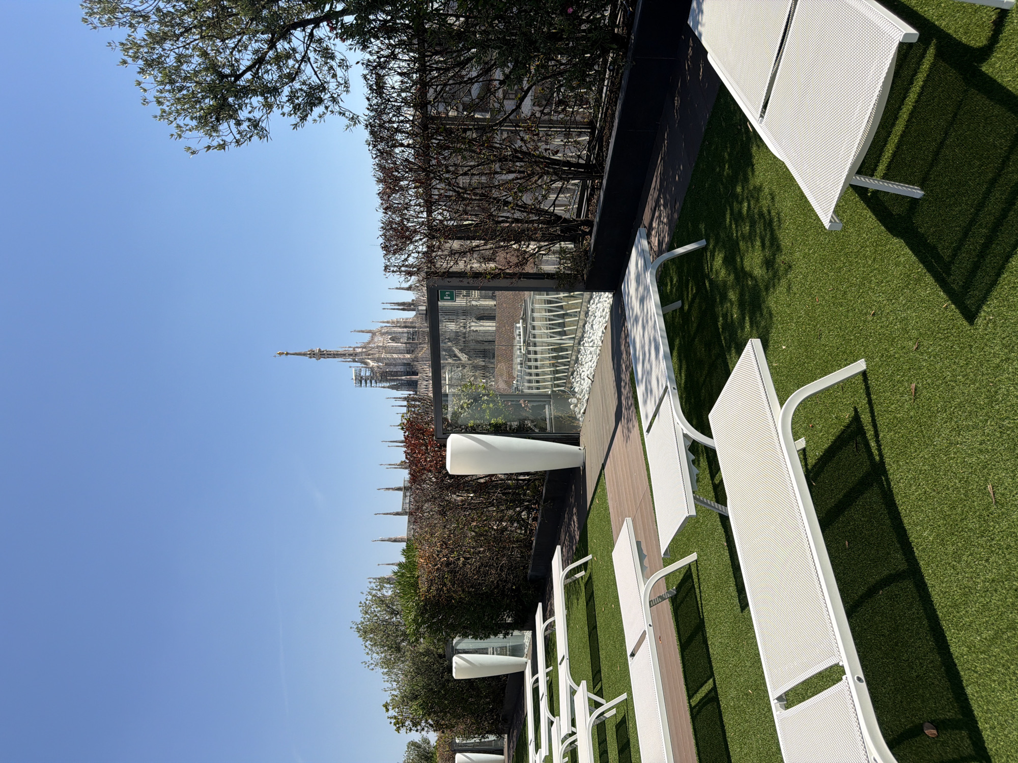The image is taken from a rooftop terrace in Milan, Italy, offering a panoramic view of the city skyline. In the foreground, several white sun loungers are arranged on a vibrant green artificial lawn. The loungers are empty and appear to be unoccupied. A wooden deck area is visible behind the loungers.
The mid-ground features a row of tall, white planters and lush green hedges, creating a sense of privacy and dividing the terrace space. Behind the hedges, a modern glass barrier is present, offering unobstructed views of the cityscape.
Dominating the background is the majestic Duomo di Milano (Milan Cathedral), a famous landmark of the city, with its intricate Gothic architecture and spires. Construction or restoration scaffolding is visible on parts of the cathedral, suggesting ongoing maintenance. The sky above is a clear, bright blue. The overall scene depicts a serene and upscale outdoor space with a stunning urban backdrop, likely a hotel rooftop or a private residence. There are no people visible in the image.
