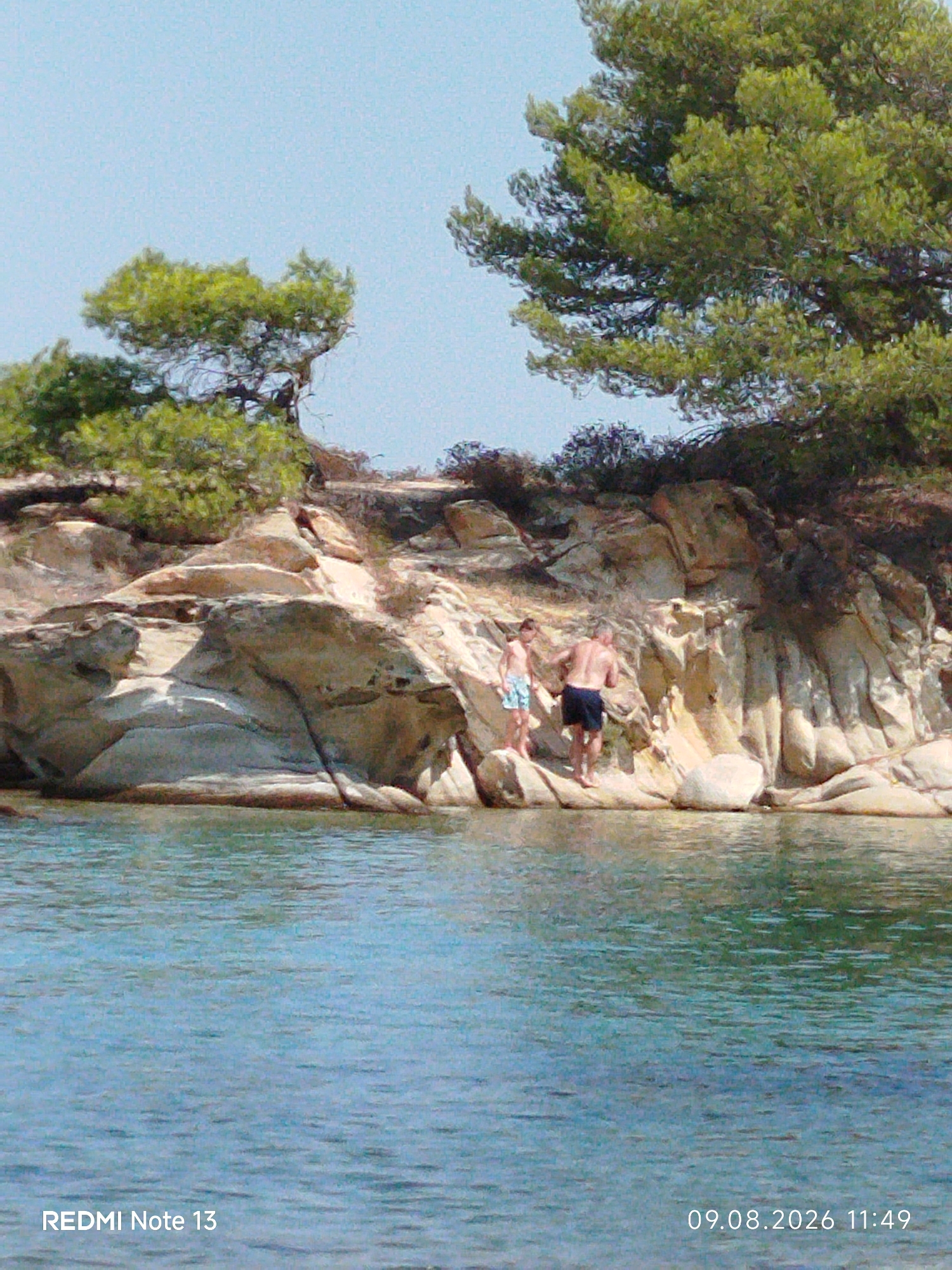 This image depicts a scenic view of the coastline in Sithonia Municipal Unit, Greece. Two individuals, an adult male and a boy, are standing on a rocky outcrop near the water. The man is wearing dark blue swim shorts, and the boy is wearing patterned swim shorts. The terrain is characterized by weathered rock formations and lush green vegetation, including pine trees. The water in the foreground is a clear turquoise color, reflecting the bright sky. The overall scene evokes a sense of a warm, sunny day at a coastal destination. The timestamp at the bottom right of the image indicates that the photo was taken on August 9, 2026, at 11:49 AM. The text "REDMI Note 13" is also visible at the bottom left, indicating the device used to capture the image.