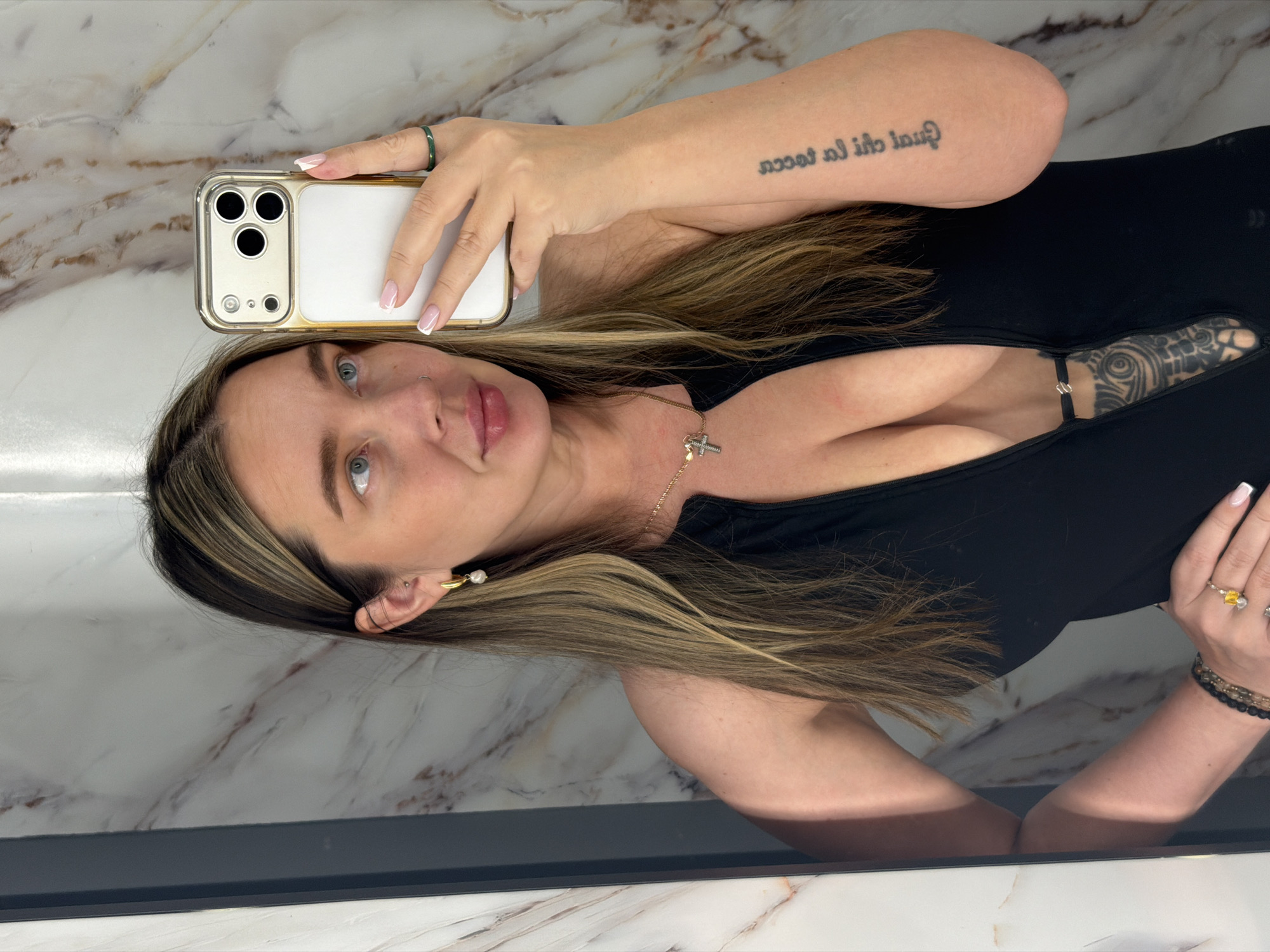 A woman with fair skin and light brown hair with blonde highlights is taking a selfie in what appears to be a bathroom. She is holding a white and gold iPhone in her left hand, which has manicured nails with white tips, and is wearing a green ring on her index finger. Her right hand, adorned with a ring with a yellow stone and bracelets made of beads, is visible near her chest. She is wearing a black top with a low neckline, revealing a tattoo on her chest. A gold necklace with a cross pendant is visible around her neck, and she has an earring with a pearl. Her hair is parted to the side. A tattoo in black ink is visible on her left forearm. The background is a marble wall and a dark-colored frame, possibly a mirror. There is no evidence of Moscow, Russia in the picture.