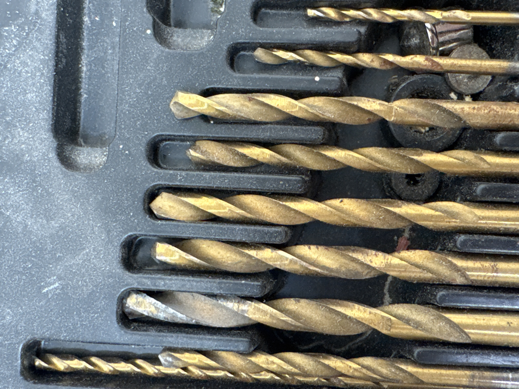 This is a close-up, overhead shot of a set of drill bits stored in a black plastic case. The drill bits are made of a golden-colored metal, likely high-speed steel, and have a twisted, fluted design. They are of varying sizes and are arranged in individual slots within the case. The case itself appears to be made of black plastic with a textured surface. The lighting suggests an outdoor environment, possibly under daylight. There are no people visible in the image. The scene depicts a collection of tools in their storage container. The location of Яромінскі сельскі Савет, Belarus is irrelevant to the visual content of the image.