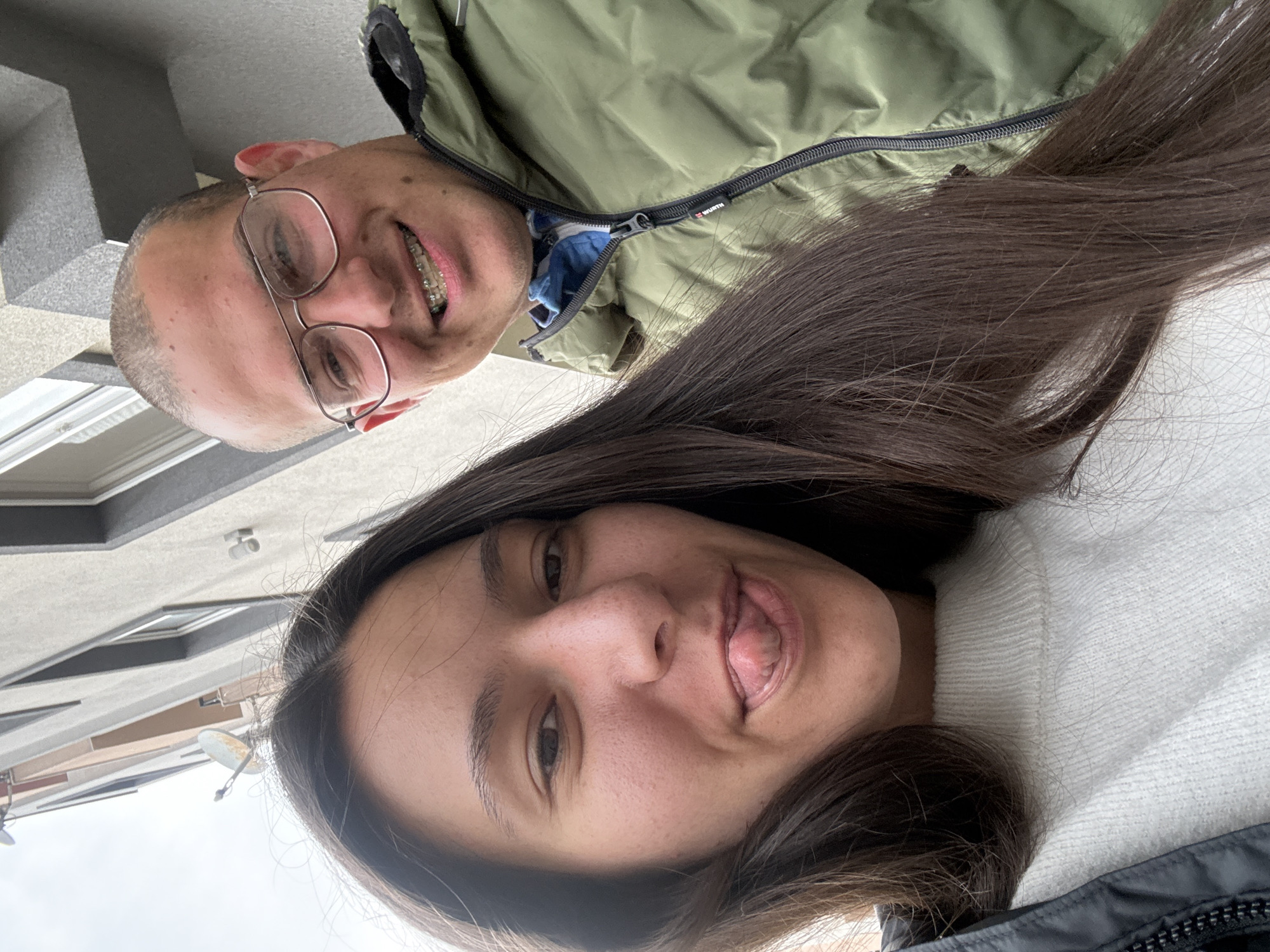 Two people are posing for a selfie outdoors. A man with short hair and glasses is wearing braces and an olive green jacket. A woman with long dark hair is wearing a white sweater and black jacket and sticking out her tongue. The background shows a building and cloudy sky. The location could not be determined to be Varna, Bulgaria.
