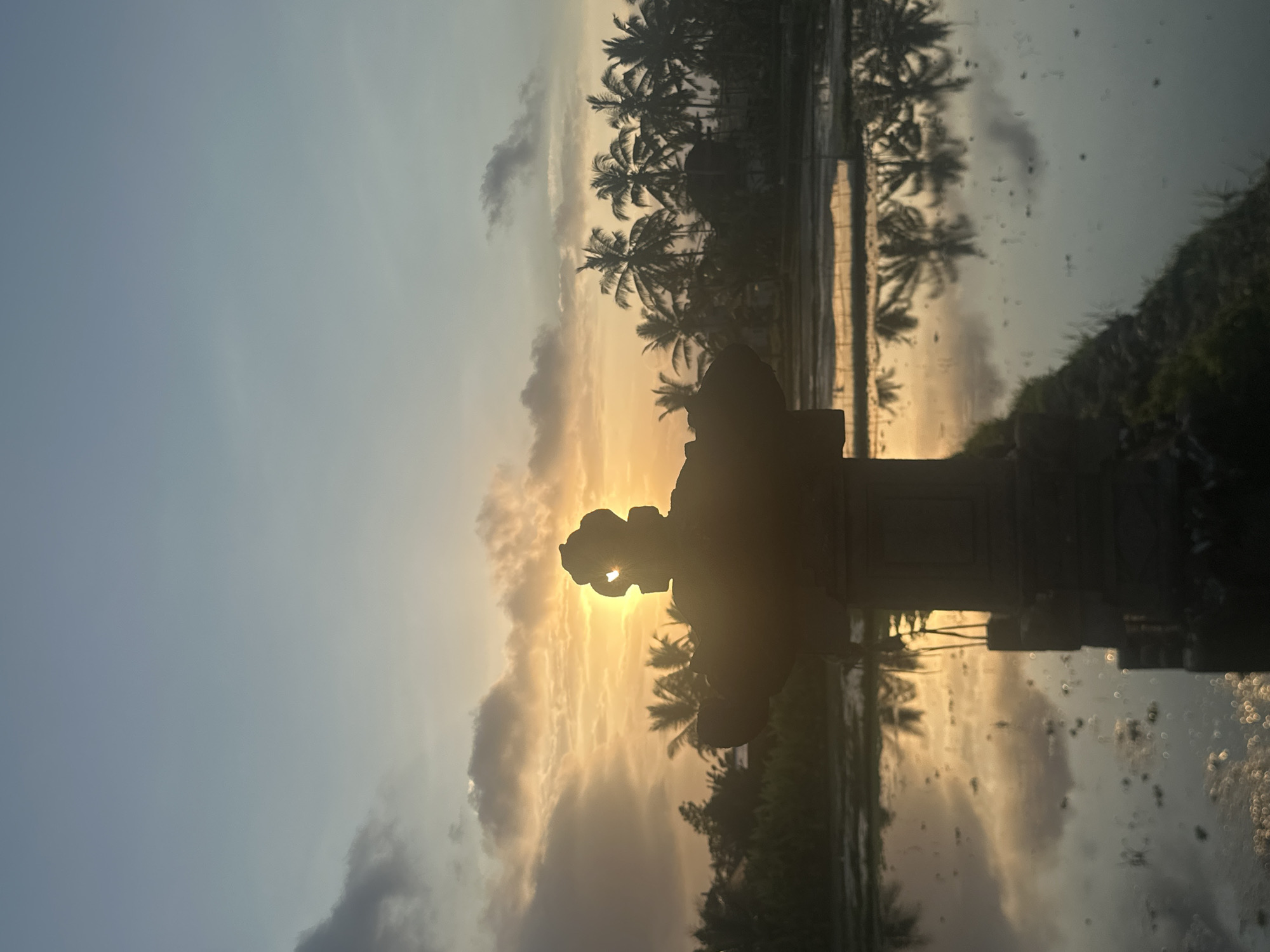 An atmospheric, silhouetted sunset is captured over a flooded rice paddy in Ubud, Indonesia. The sun, a bright orb of orange and yellow, is partially obscured by dark, textured clouds and peeks through a carved opening in a stone sculpture in the foreground. The sculpture, with intricate details and a wide base, stands prominently in the center of the frame, casting a long shadow.
The tranquil water of the paddy reflects the vibrant sky, the silhouettes of palm trees, and the surrounding vegetation. The trees, a mix of tall and shorter varieties, form a dense line along the horizon, their fronds distinct against the soft hues of the sky. The overall impression is one of serene, natural beauty, amplified by the warm glow of the setting sun. The scene suggests a rural or agricultural setting, typical of the landscape found in Ubud.