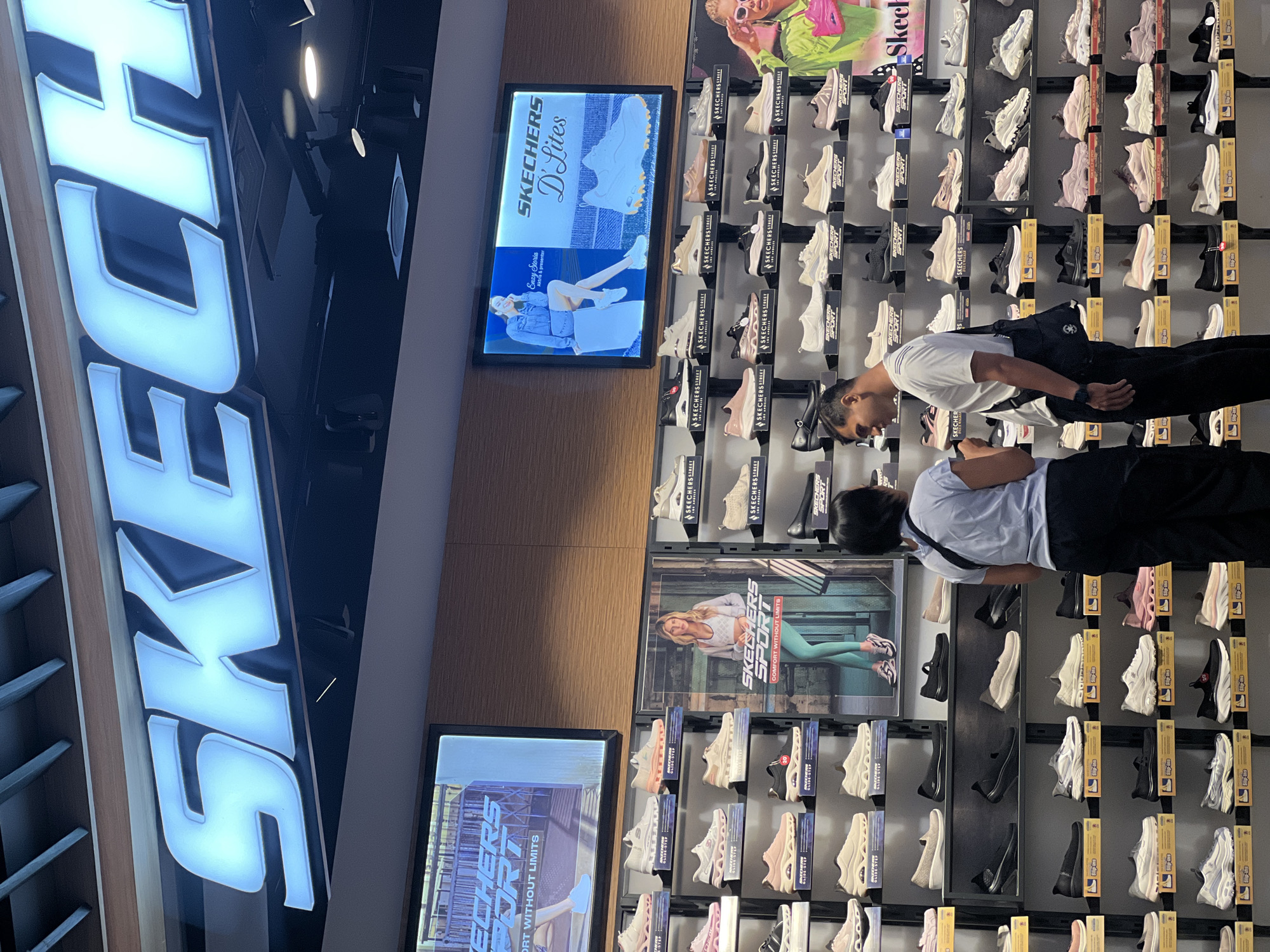 The image shows the interior of a Skechers store in Denpasar, Indonesia. The store is well-lit and features a large display of Skechers shoes on shelves lining the walls. Above the shelves, illuminated signs display the Skechers logo and advertisements for Skechers products. Two individuals, a man and a woman, are standing in front of a shoe display, appearing to be shopping. The man is wearing a white t-shirt and black pants, while the woman is wearing a blue t-shirt and black pants. The overall atmosphere suggests a typical retail environment.