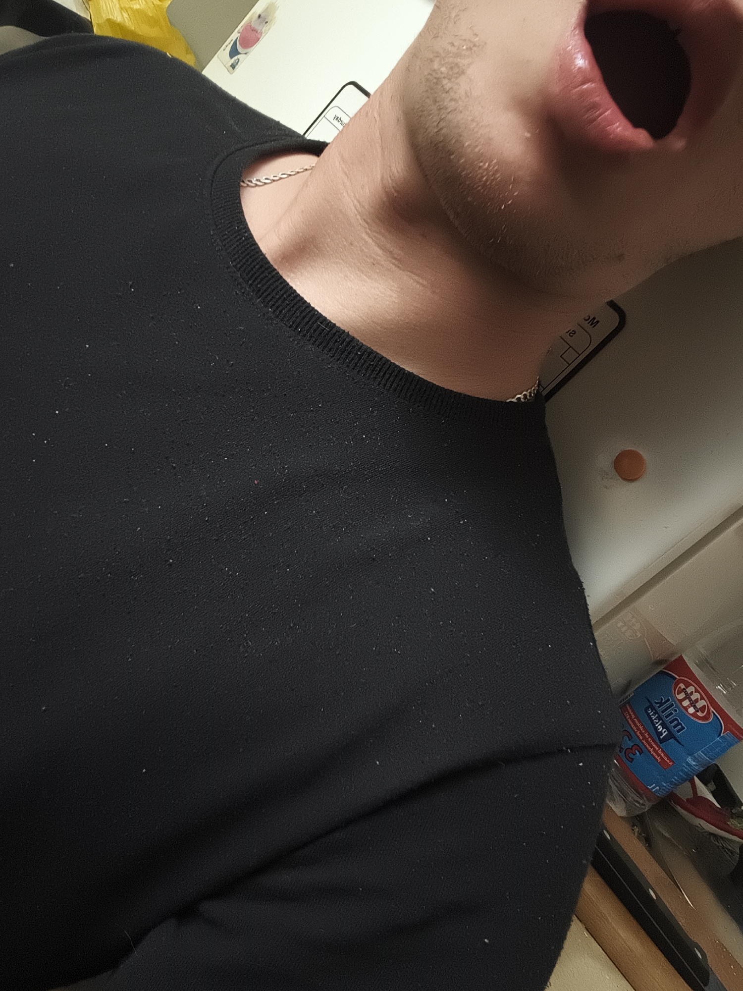 A close-up, slightly angled shot shows a person wearing a black crew-neck shirt with a silver chain visible around their neck. Their mouth is open, revealing the interior. The person's face is close to the side of what appears to be a white refrigerator or cabinet. A red and blue carton labeled "Slim" is visible near the lower right corner, amidst other items in a partial view of a cluttered area. A small orange circular object is also on the surface near the carton. There is no specific information in the image to determine its precise location in the city of Nowy Tomyśl, Poland.